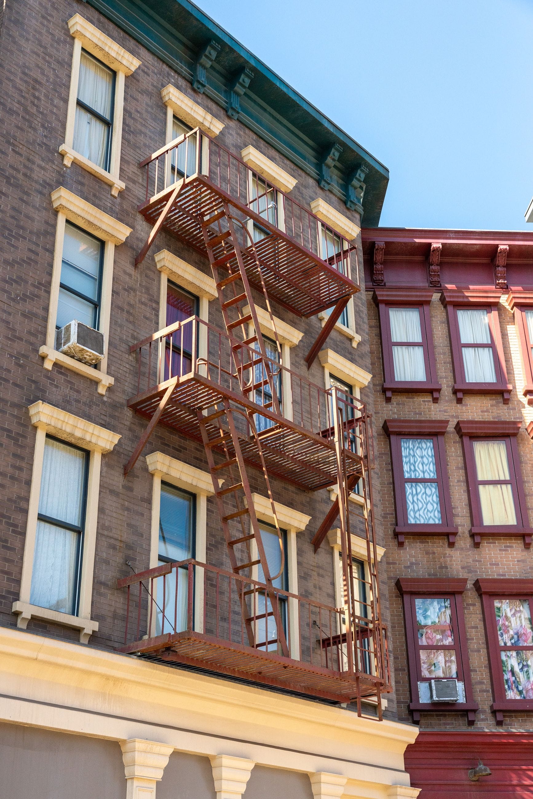 Brown brick building with a rusty fire escape next to a reddish brick building under a blue sky.
