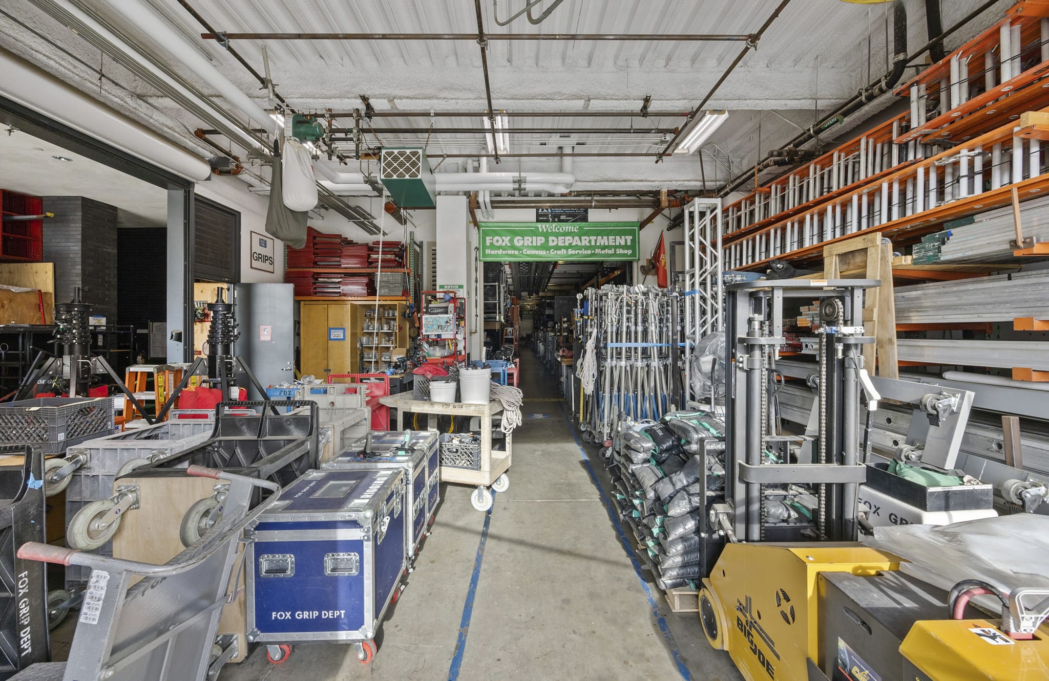 A long aisle in a film equipment warehouse with cases, stands, ladders, a forklift, and a "FOX GRIP DEPARTMENT" sign.