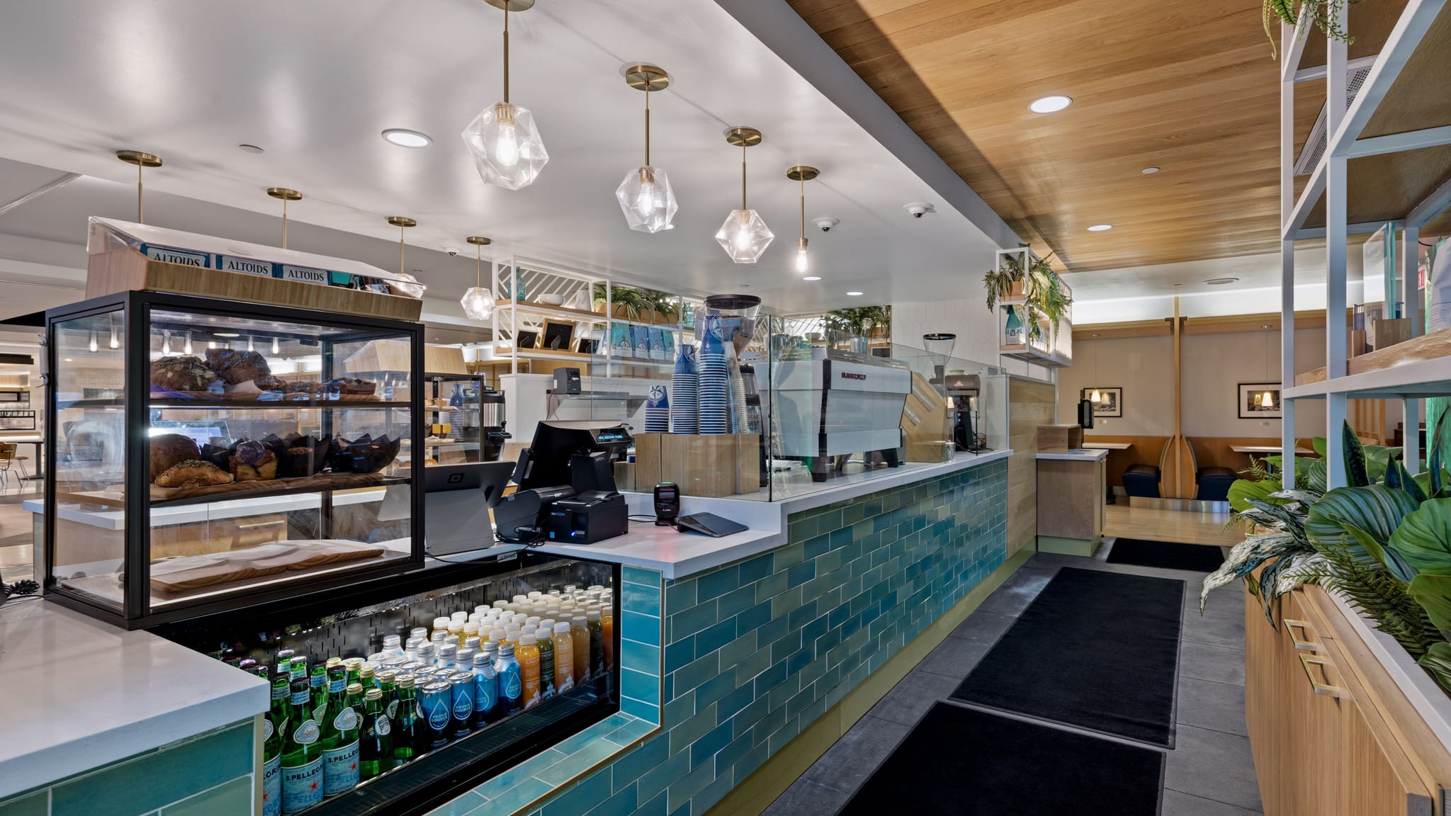 A modern cafe interior with a turquoise tiled counter, a pastry display, coffee machines, and a seating area with wooden booths and plants.