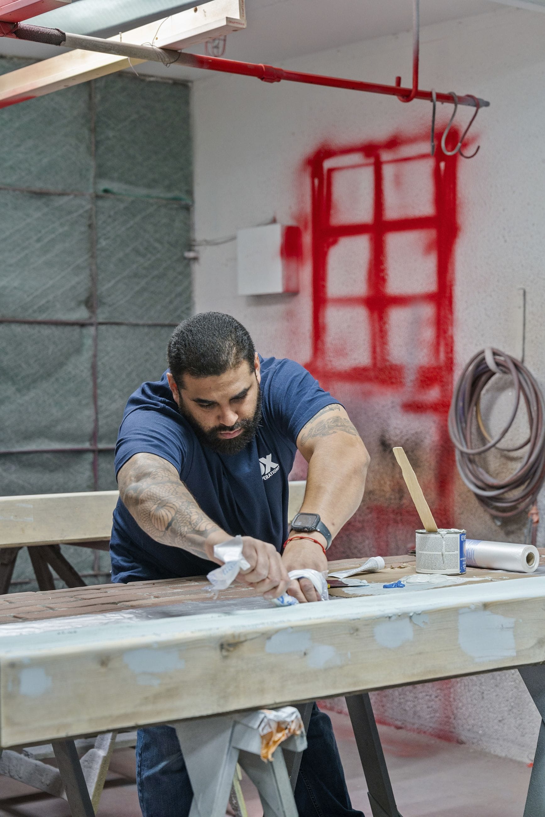 A bearded man with tattoos sands or finishes a wooden beam in a workshop.