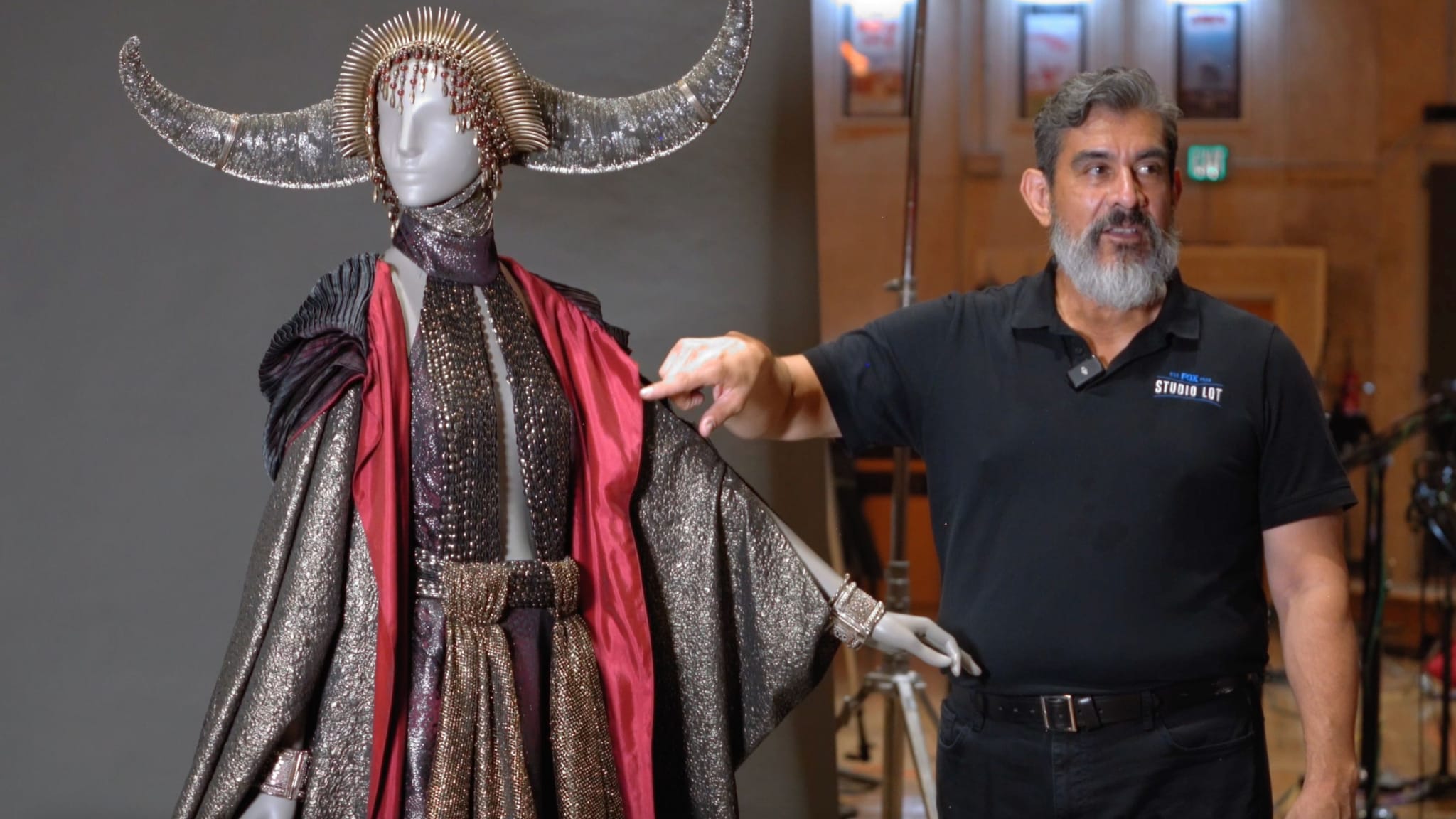 A man gestures towards a mannequin displaying an elaborate dark costume with a red lining and a large, horned headpiece.