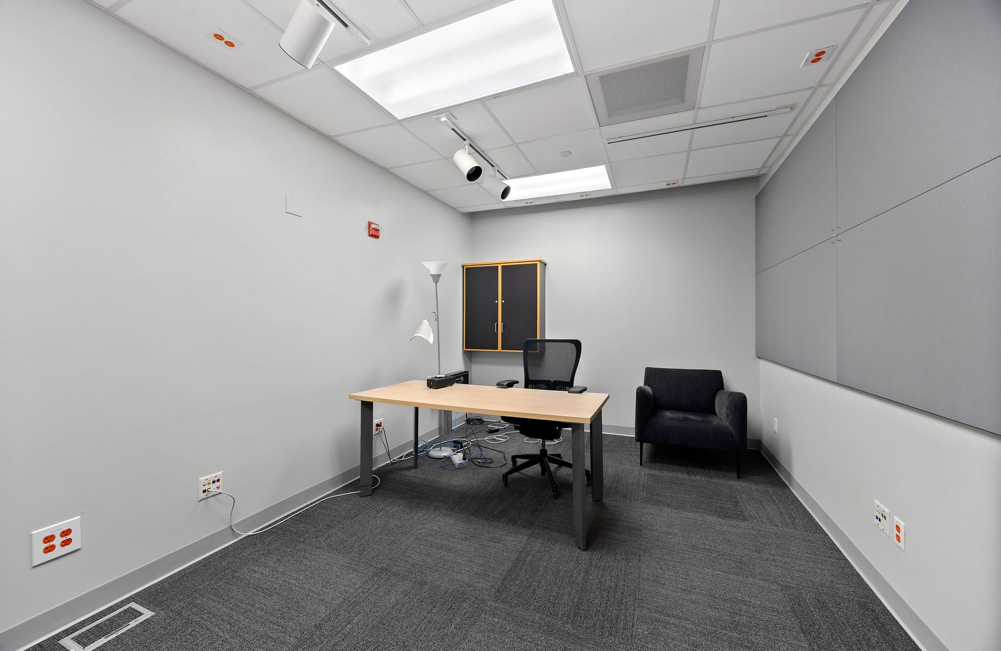 A modern office room with a light wood desk, black office chair, dark armchair, and a wood cabinet against gray walls and carpeted floor.
