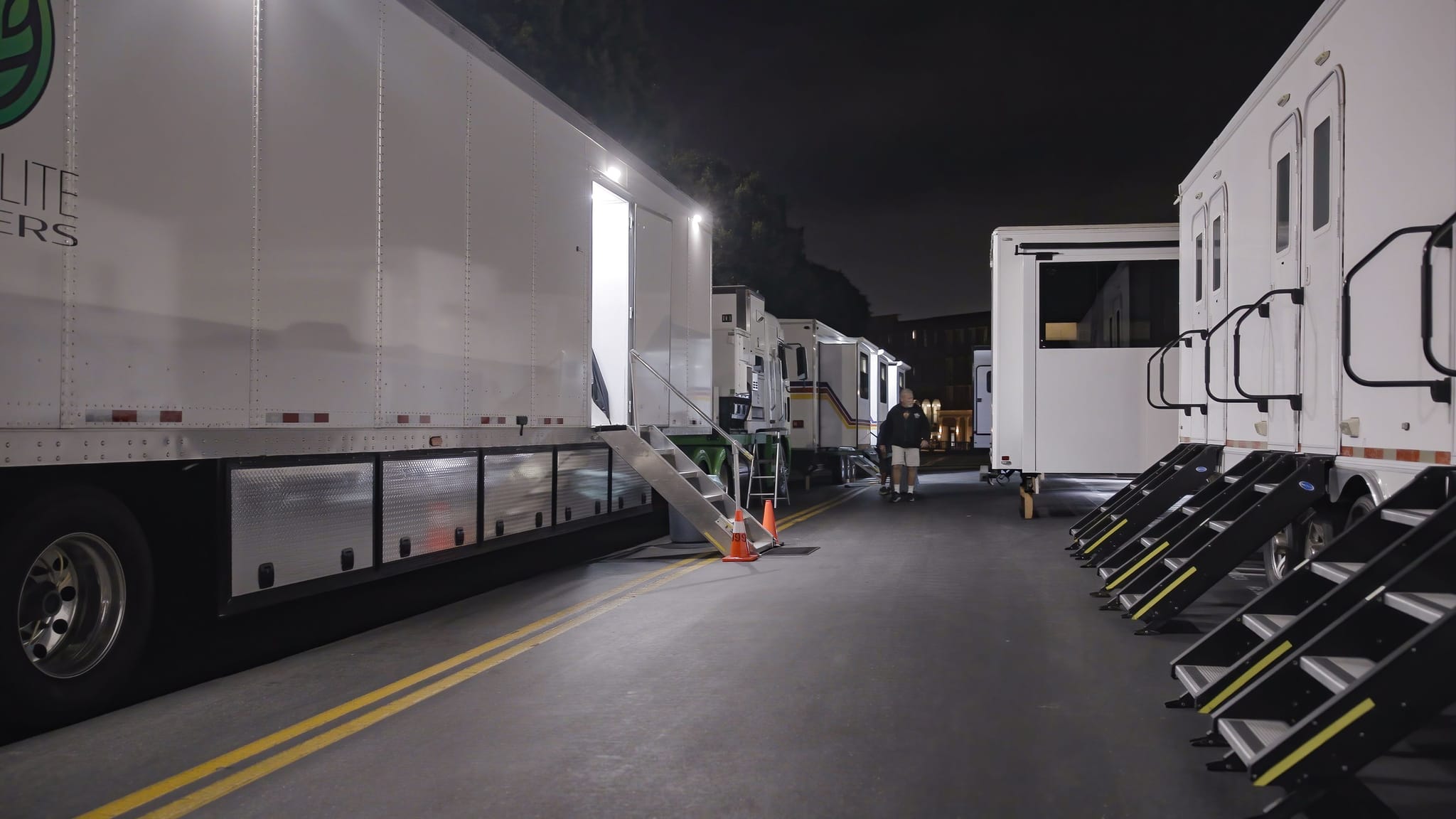 White production trailers parked on a dark lot at night, one open and brightly lit, with stairs leading to multiple others.