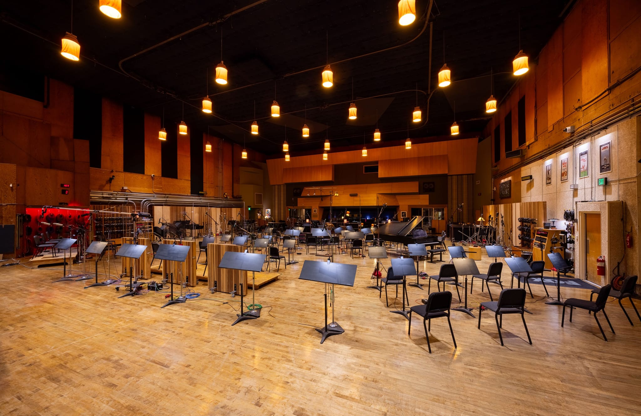A spacious recording studio with wooden floors, acoustic walls, and rows of music stands and chairs, a grand piano visible in the distance.