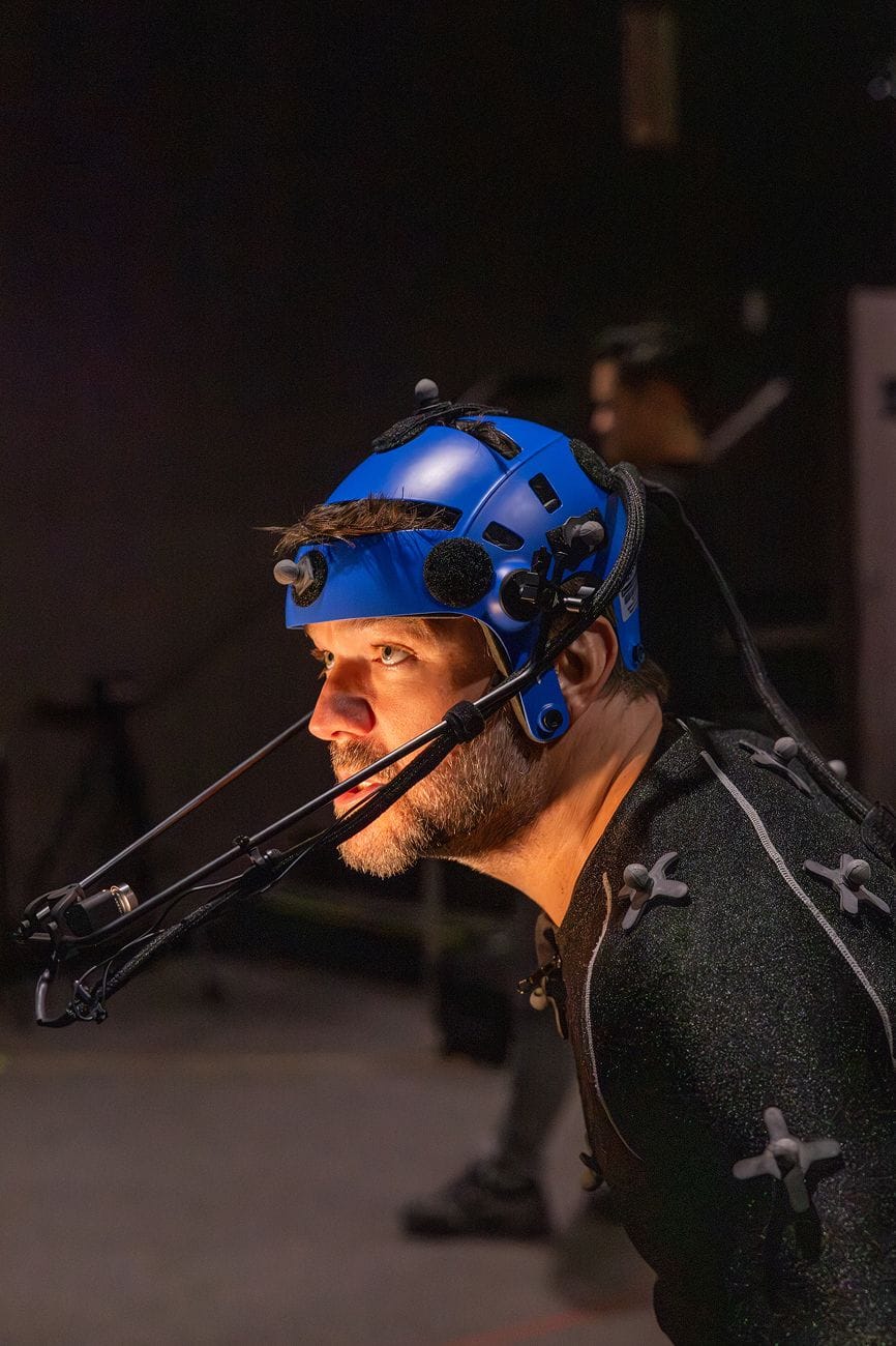 A man with a beard wears a blue motion capture helmet with a face camera rig and a tracking suit.