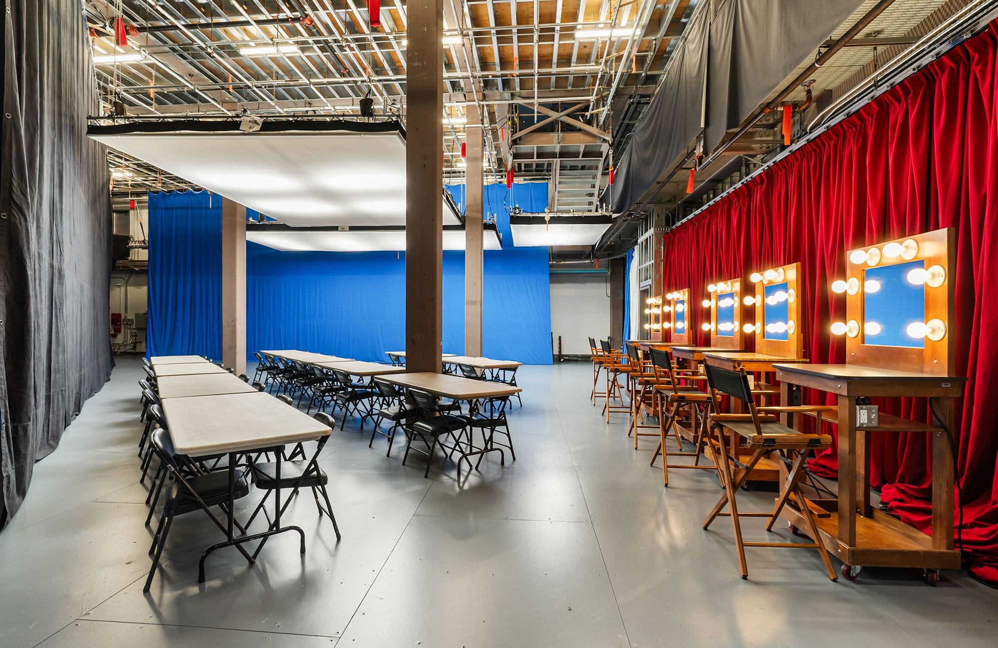 A large studio space with rows of tables, a blue screen wall, and a makeup station with lighted mirrors and red curtains.