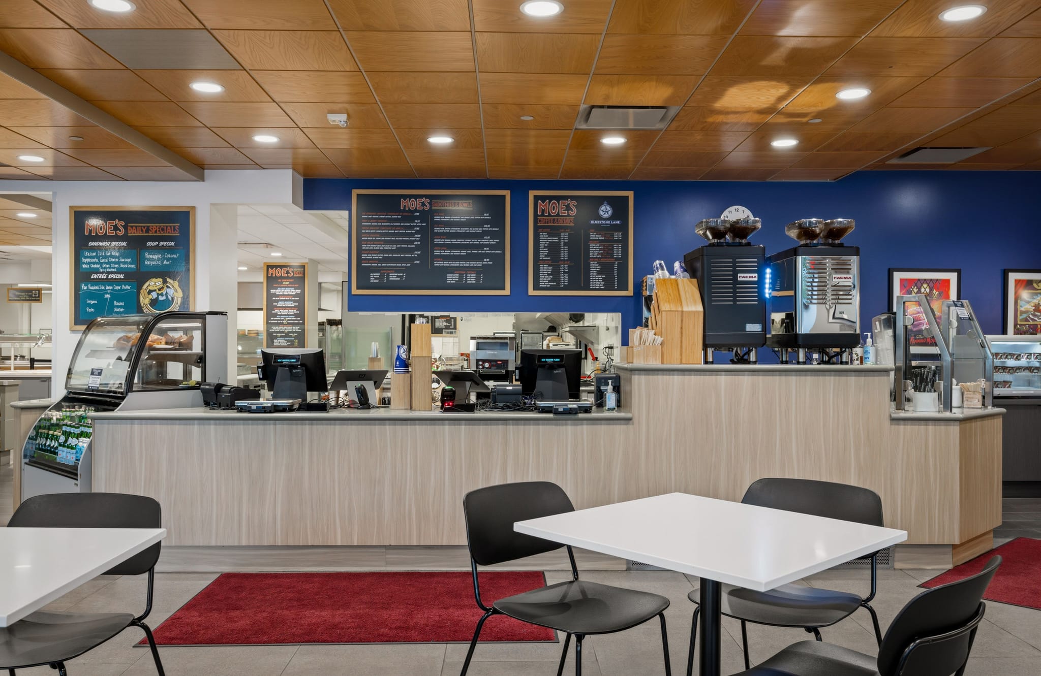 A modern cafe with a light wood-paneled counter, a blue wall with Moe's menu boards, coffee machines, and tables with chairs in the foreground.
