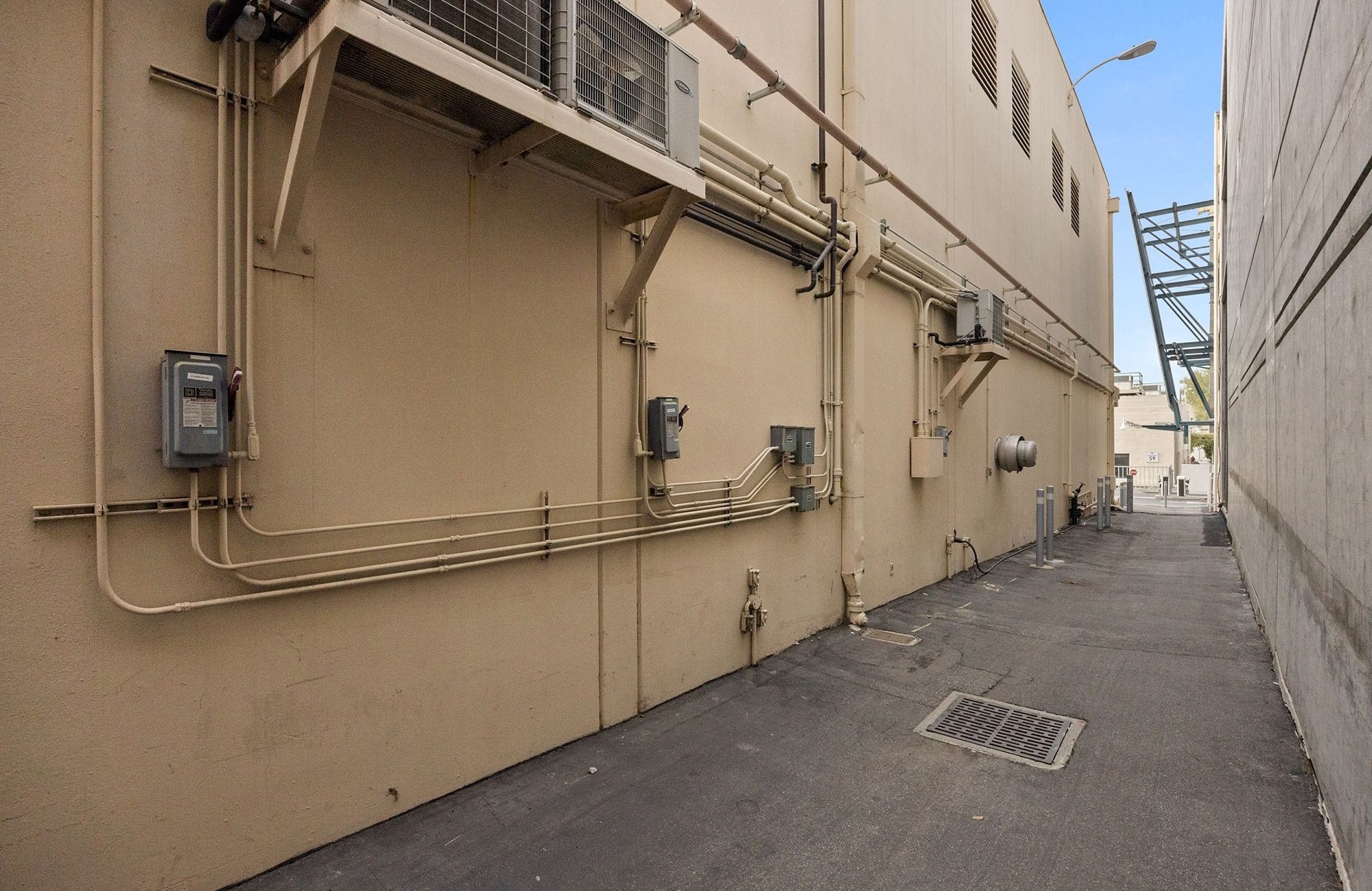 A narrow alleyway between two buildings, with a tan wall on the left showing exposed pipes and utility boxes.