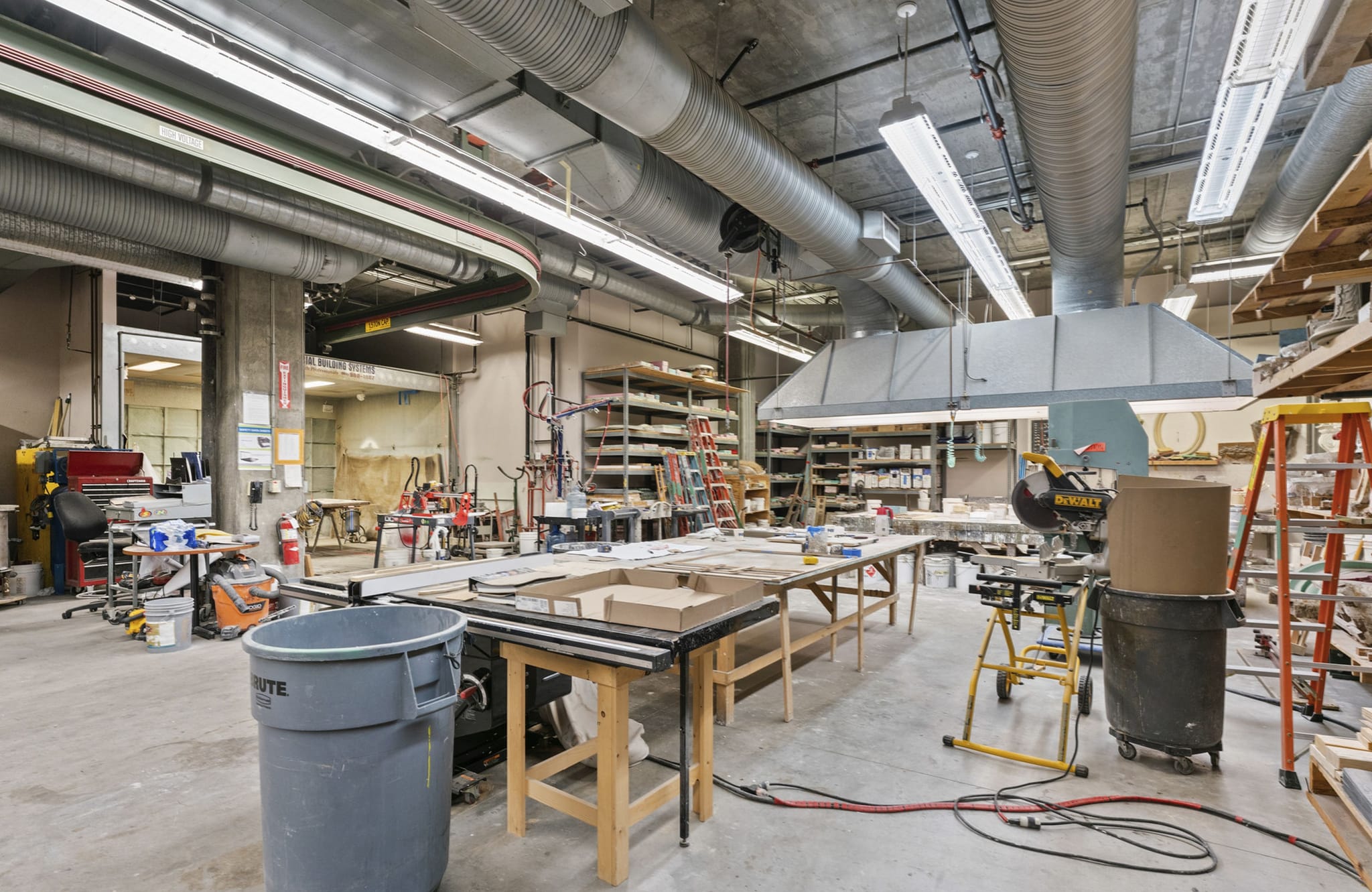 A large, well-lit workshop with workbenches, power tools, shelving, and exposed ceiling ducts.
