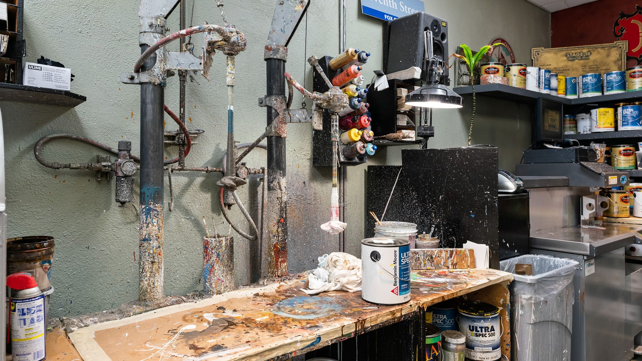 A paint-splattered paint mixing station with a tinting machine, a messy workbench, and shelves of paint cans.