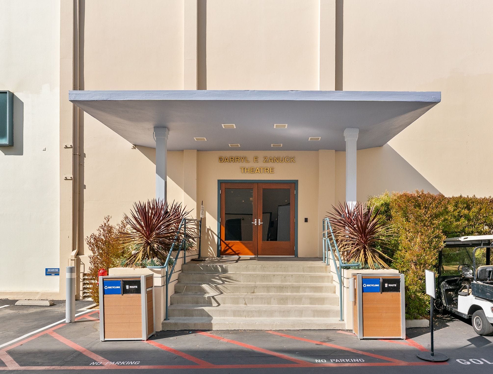 The front entrance of the Sarah E. Sangar Theatre, with concrete steps leading to double wooden doors under a light blue awning, flanked by plants and recycling bins.