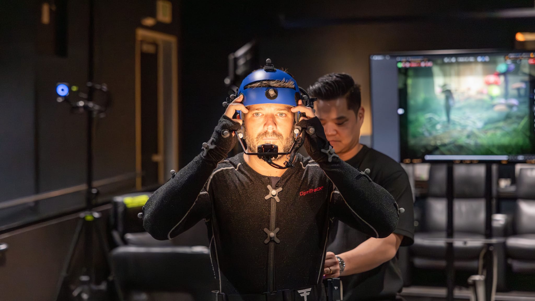 A man in a motion capture suit and helmet is adjusted by an assistant, with a virtual scene on a monitor in the background.