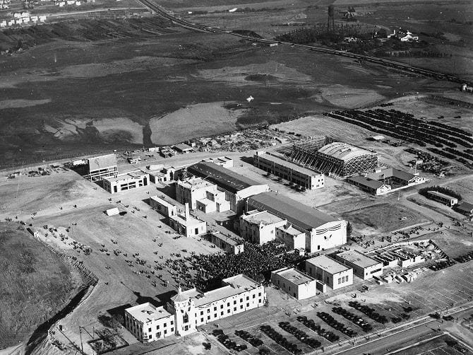 Black and white aerial view of a large industrial or studio complex with many buildings, parked cars, and a gathering crowd, bordered by marshland.