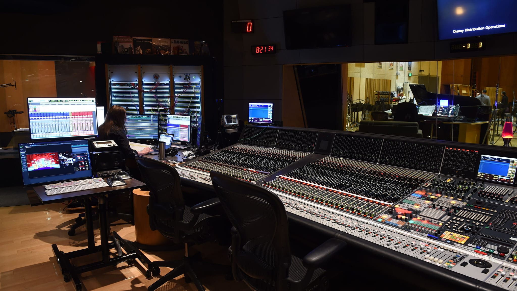 A person works at a large audio mixing console and computer monitors in a recording studio control room, with a live room visible through a window.