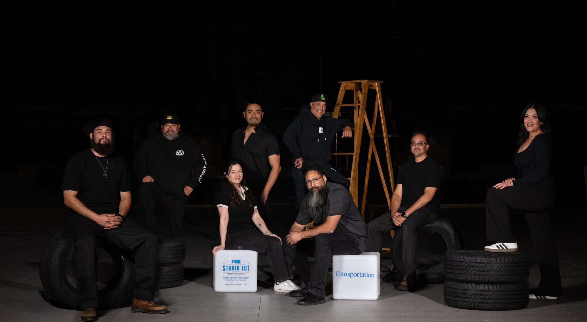 Eight people in black clothing pose with tires, a ladder, and two cubes, one labeled "PBS Studio 101" and the other "Transportation," against a dark background.