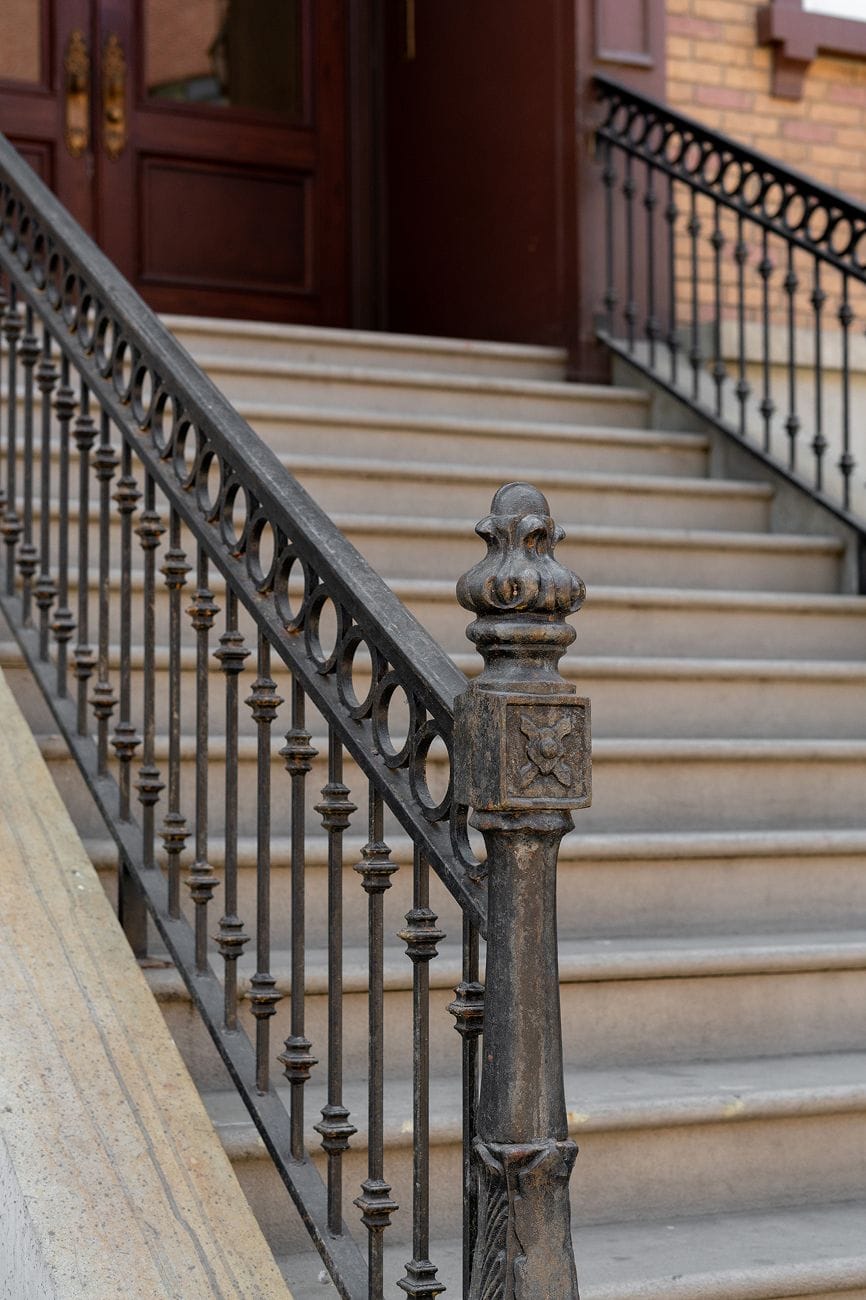An ornate dark metal railing with a decorative newel post alongside light stone steps leading to a dark brown door.