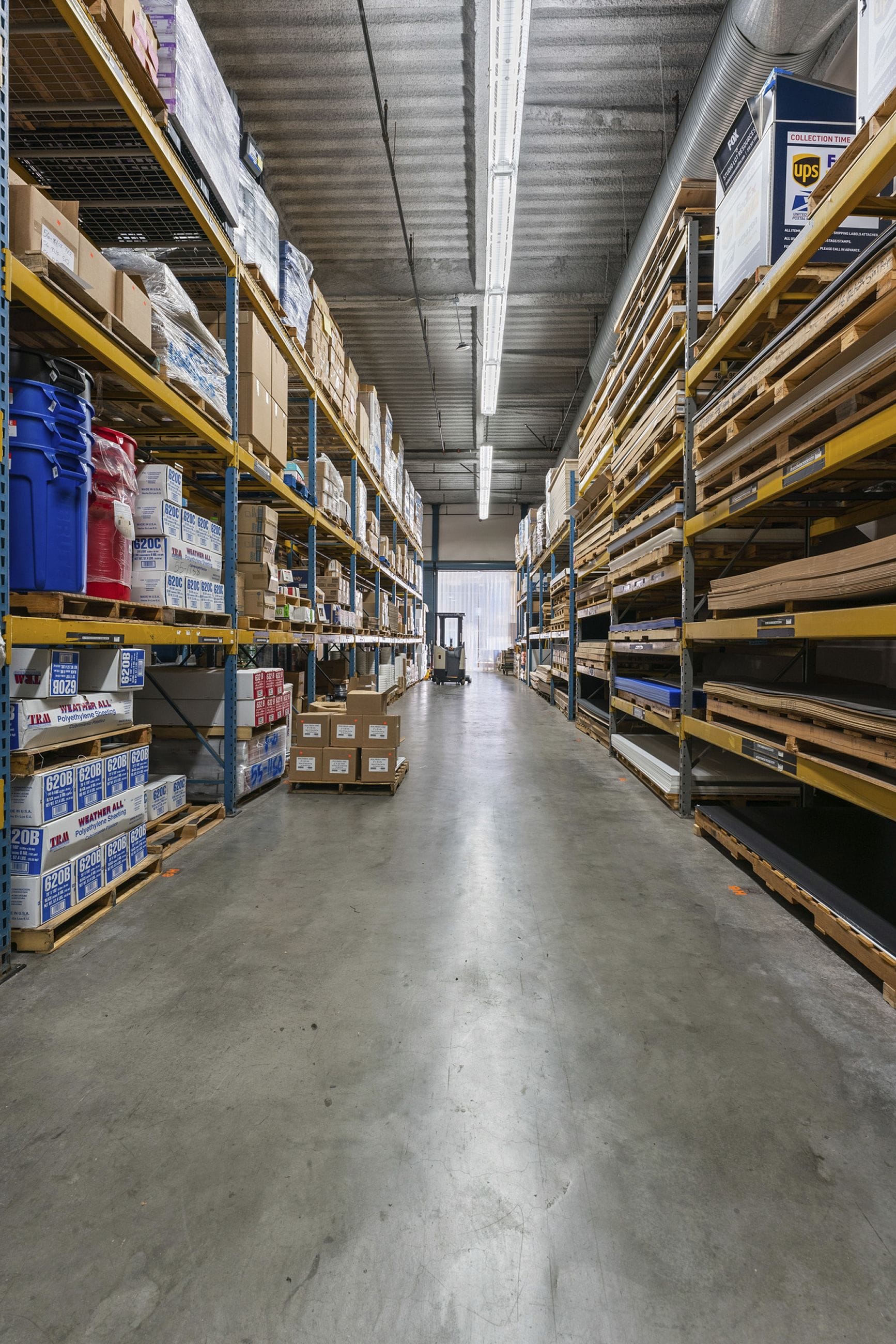 A long warehouse aisle lined with tall shelving racks filled with boxes and products, with a forklift visible in the background.