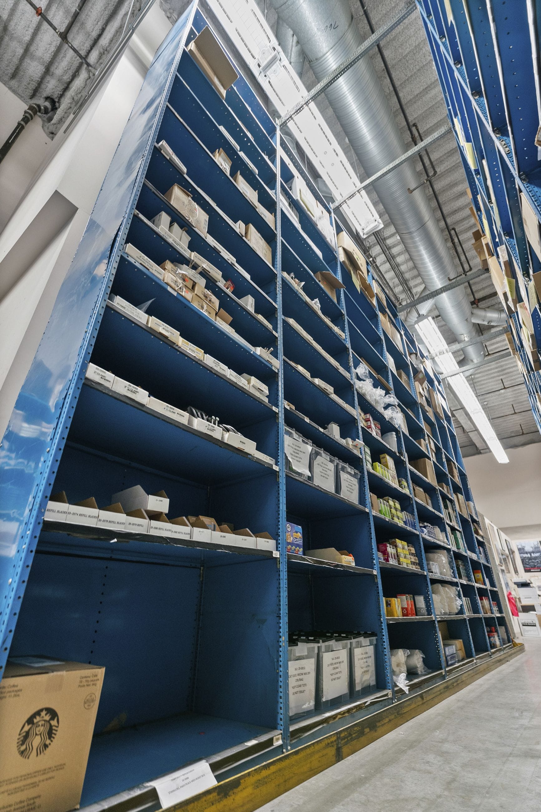 Tall blue metal mobile shelving units, filled with various items, extend towards an industrial ceiling with ducts.