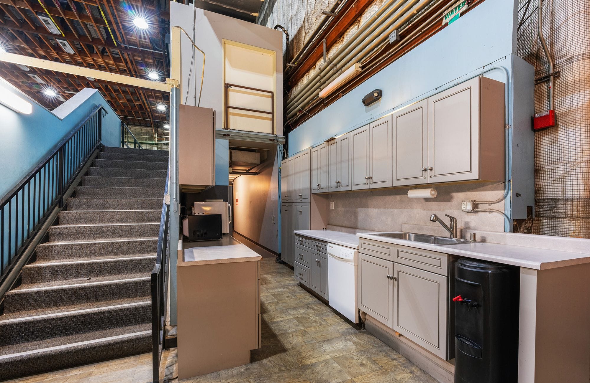 A kitchen with light gray cabinets and a sink next to a carpeted staircase in an industrial setting.