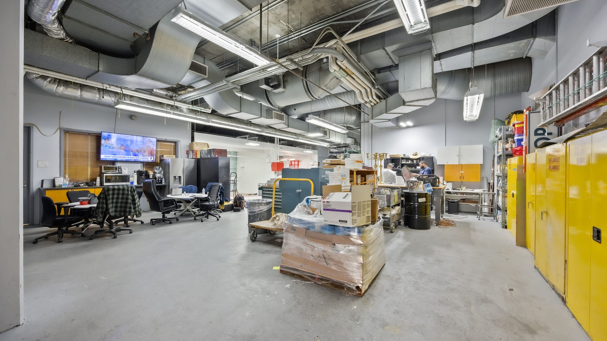 An industrial-style workshop or lab with exposed ducts, featuring desks, workbenches, and yellow storage cabinets.