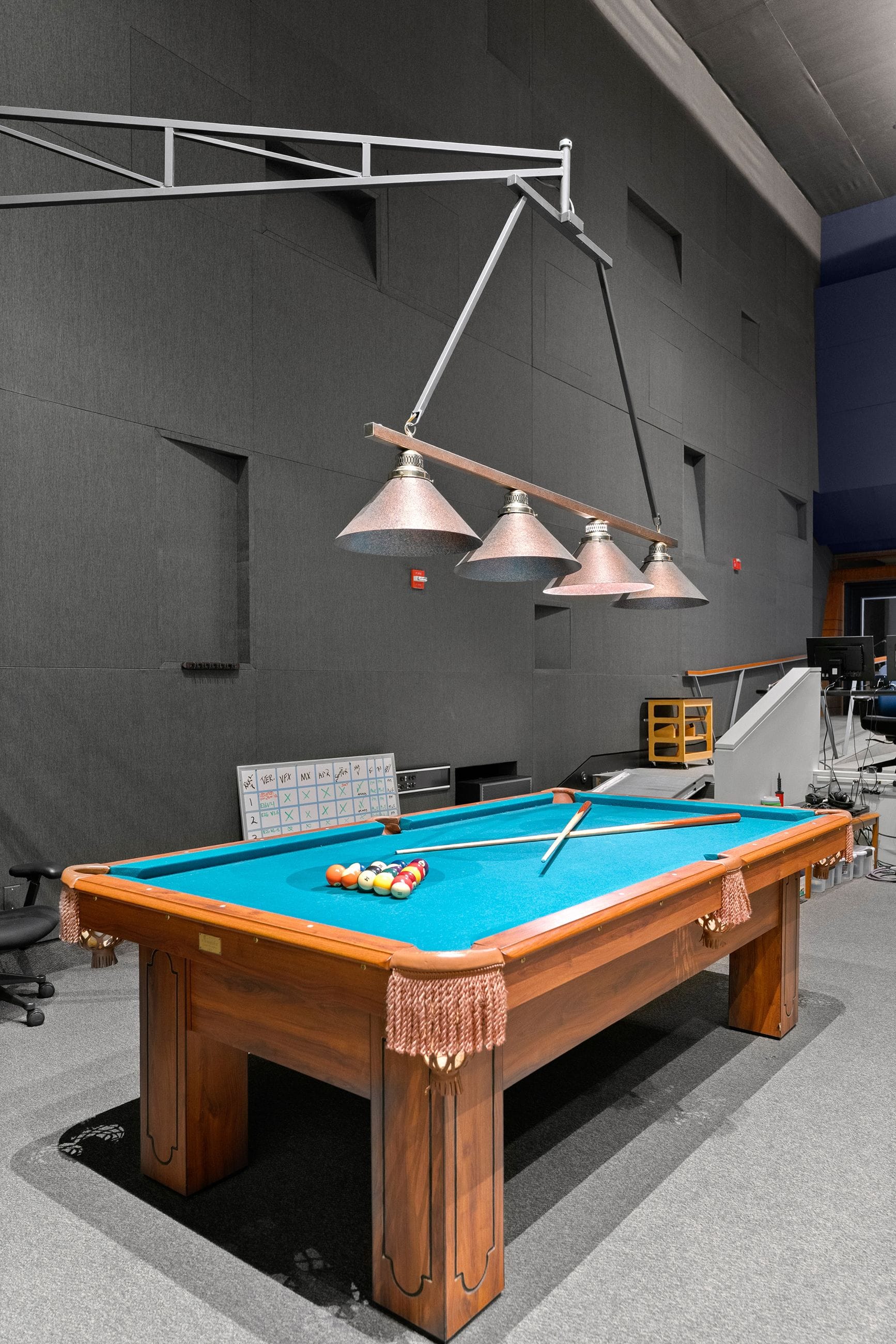A wooden pool table with blue felt, racked balls, and cues sits under a four-light pendant fixture in a large room with dark gray acoustic walls.