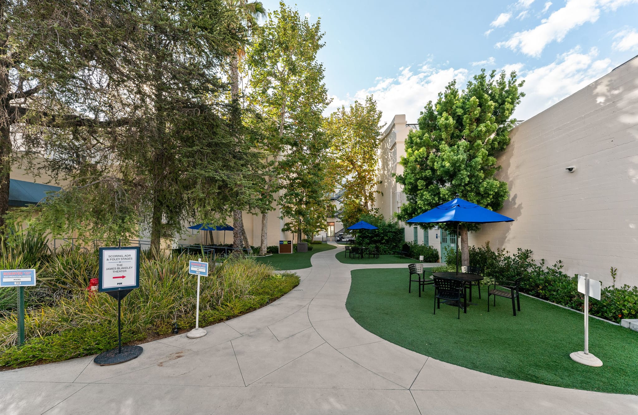A green courtyard with a winding concrete path, trees, artificial turf, and two blue patio umbrellas with seating.
