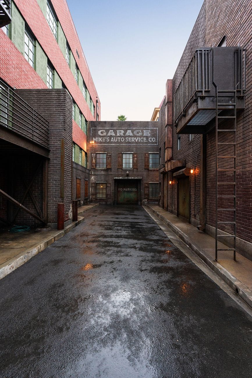 A wet asphalt alleyway flanked by brick buildings leads to "Mike's Auto Service" garage, with a "No Parking" sign on its door.