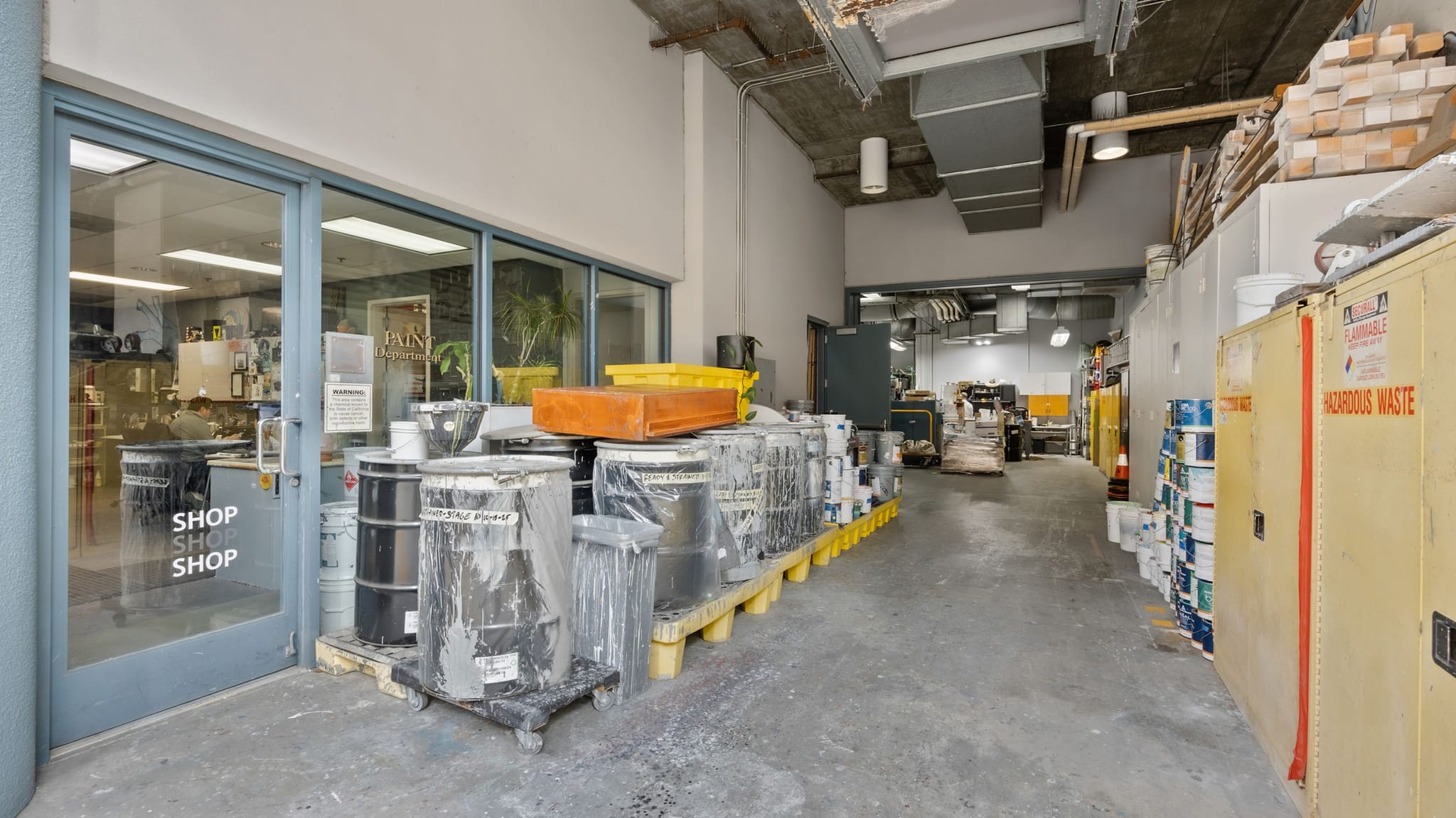 An industrial hallway with drums, paint containers, and a hazardous waste cabinet, next to a glass shop entrance.