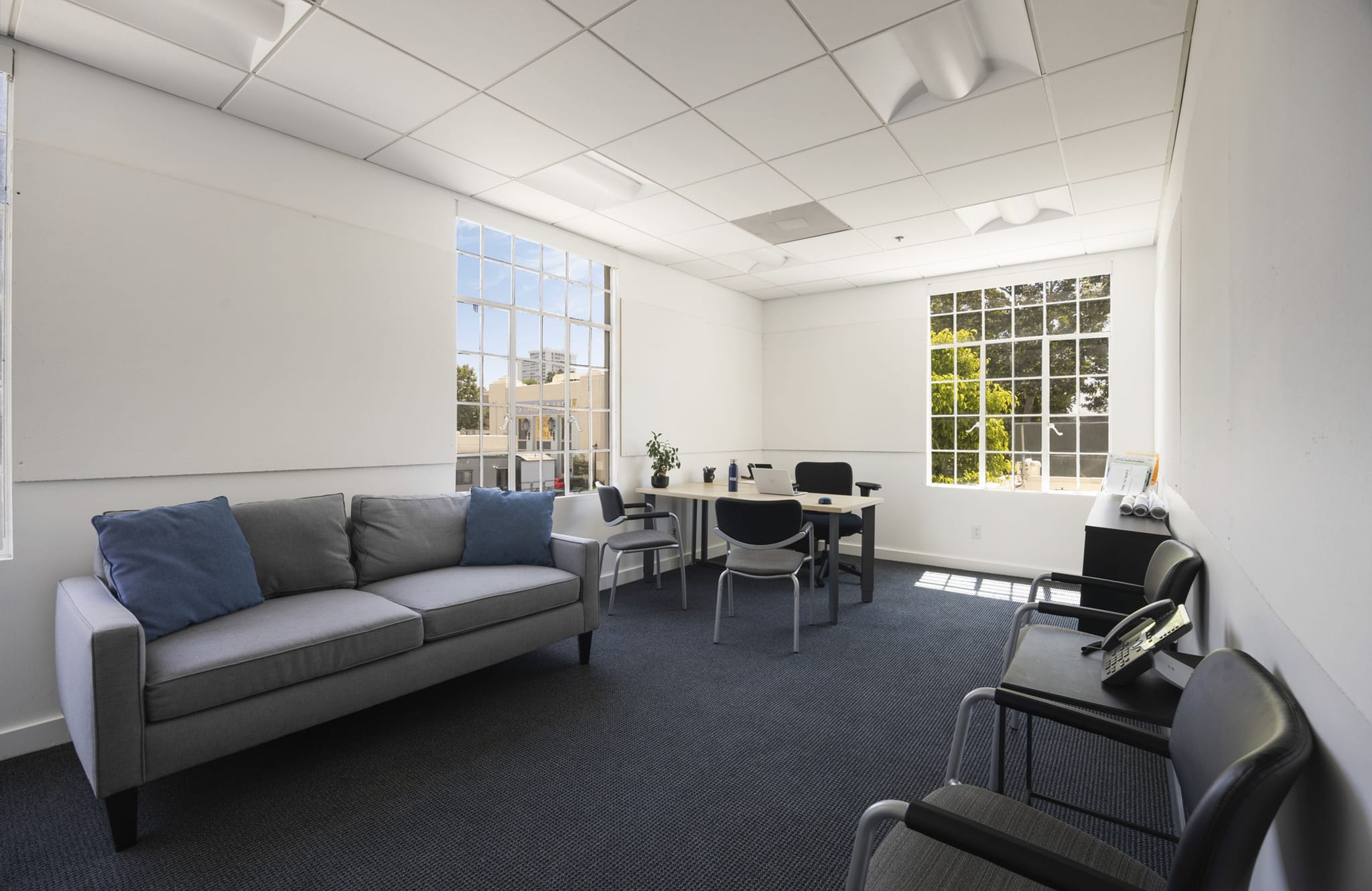 A bright, modern office space with a grey sofa, a small meeting table with chairs, and a waiting area with black chairs, all on a blue carpet.