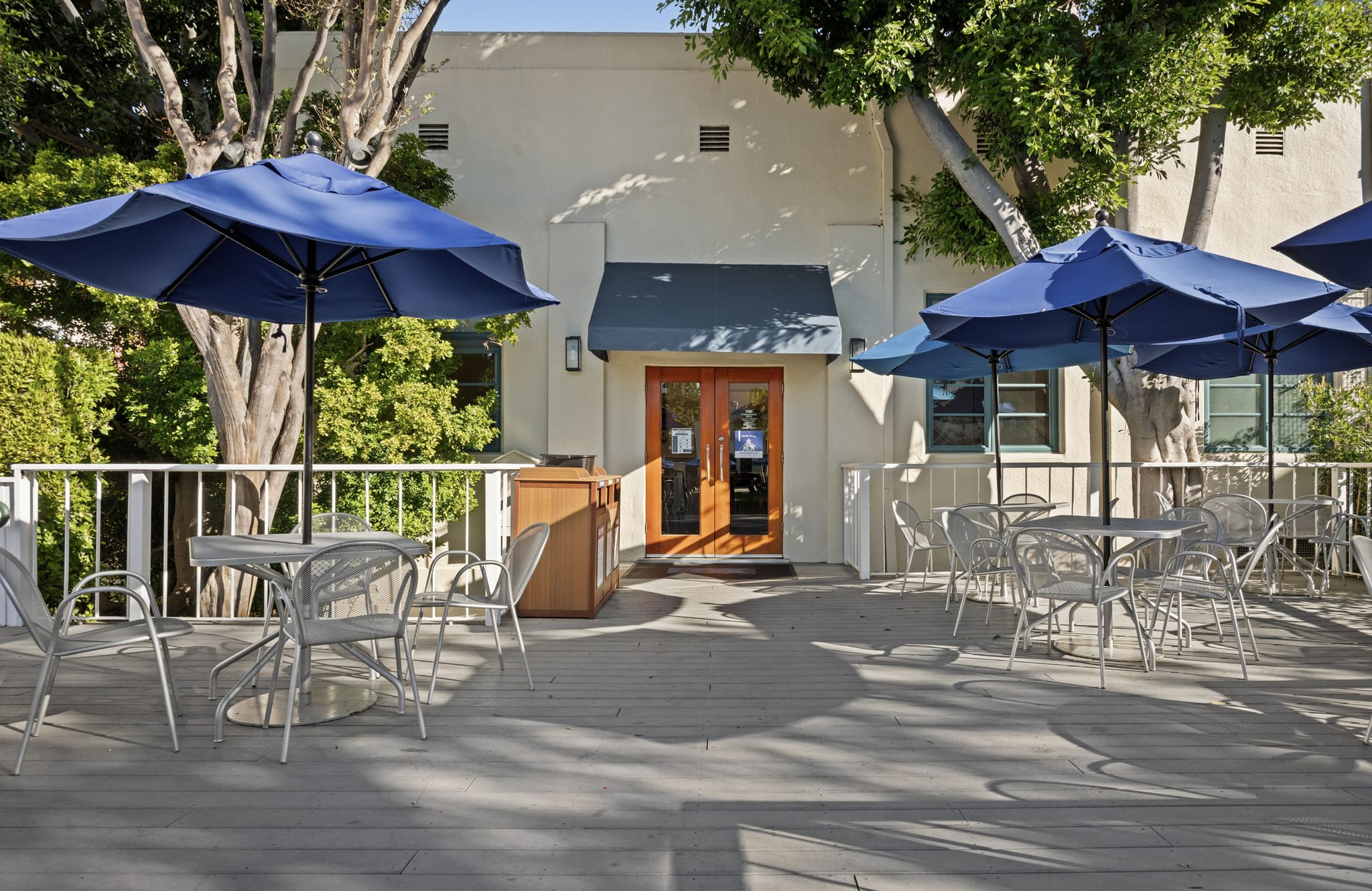Outdoor patio with blue umbrellas, tables, chairs, outside a building, with trees in the background.