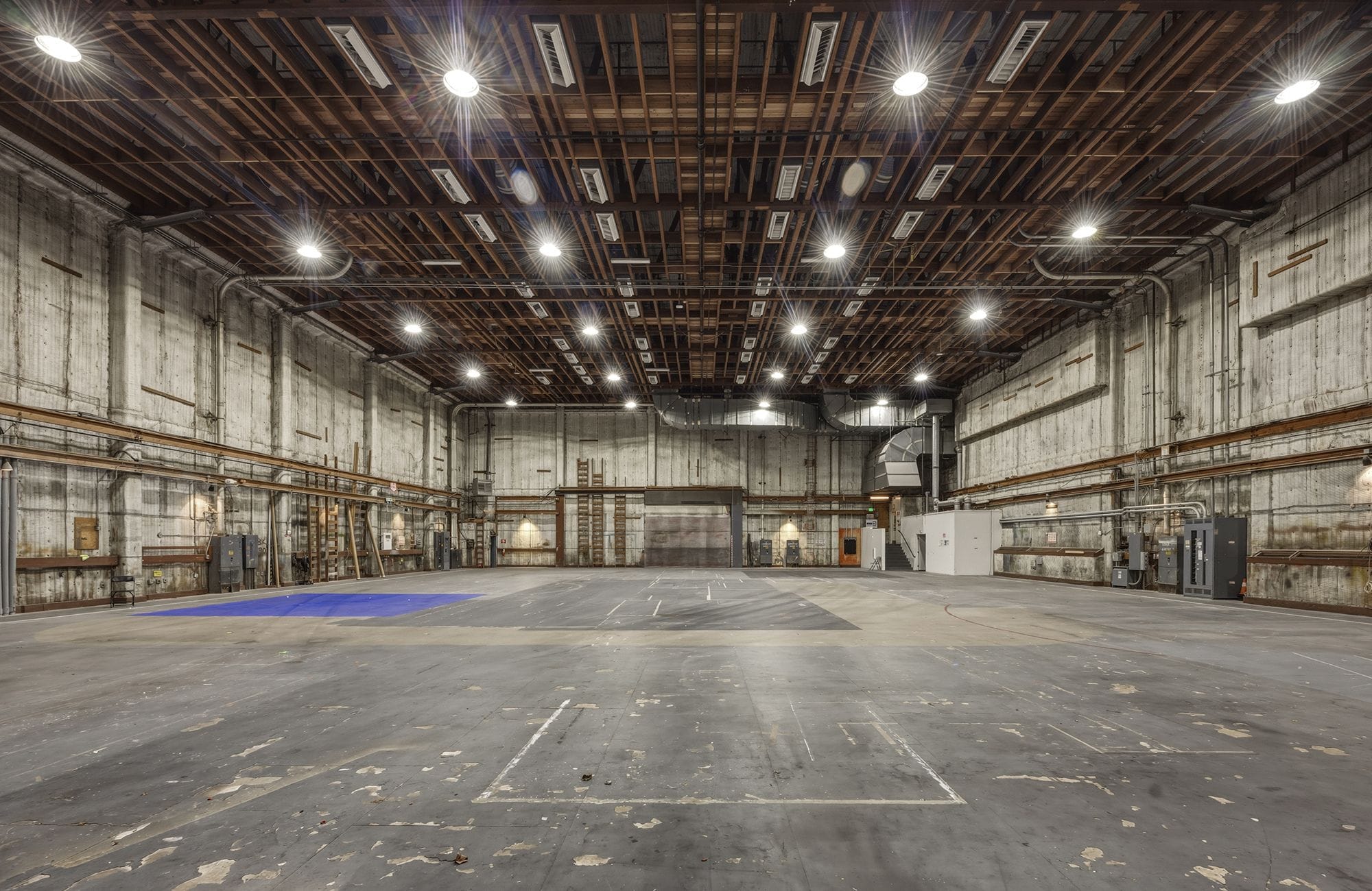 An empty, brightly lit industrial studio or warehouse with concrete walls, an exposed wooden beam ceiling, and a distressed concrete floor.