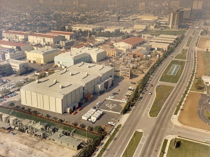Aerial view of a film studio complex with multiple soundstages, parking lots, a wide boulevard, and a hazy city in the distance.