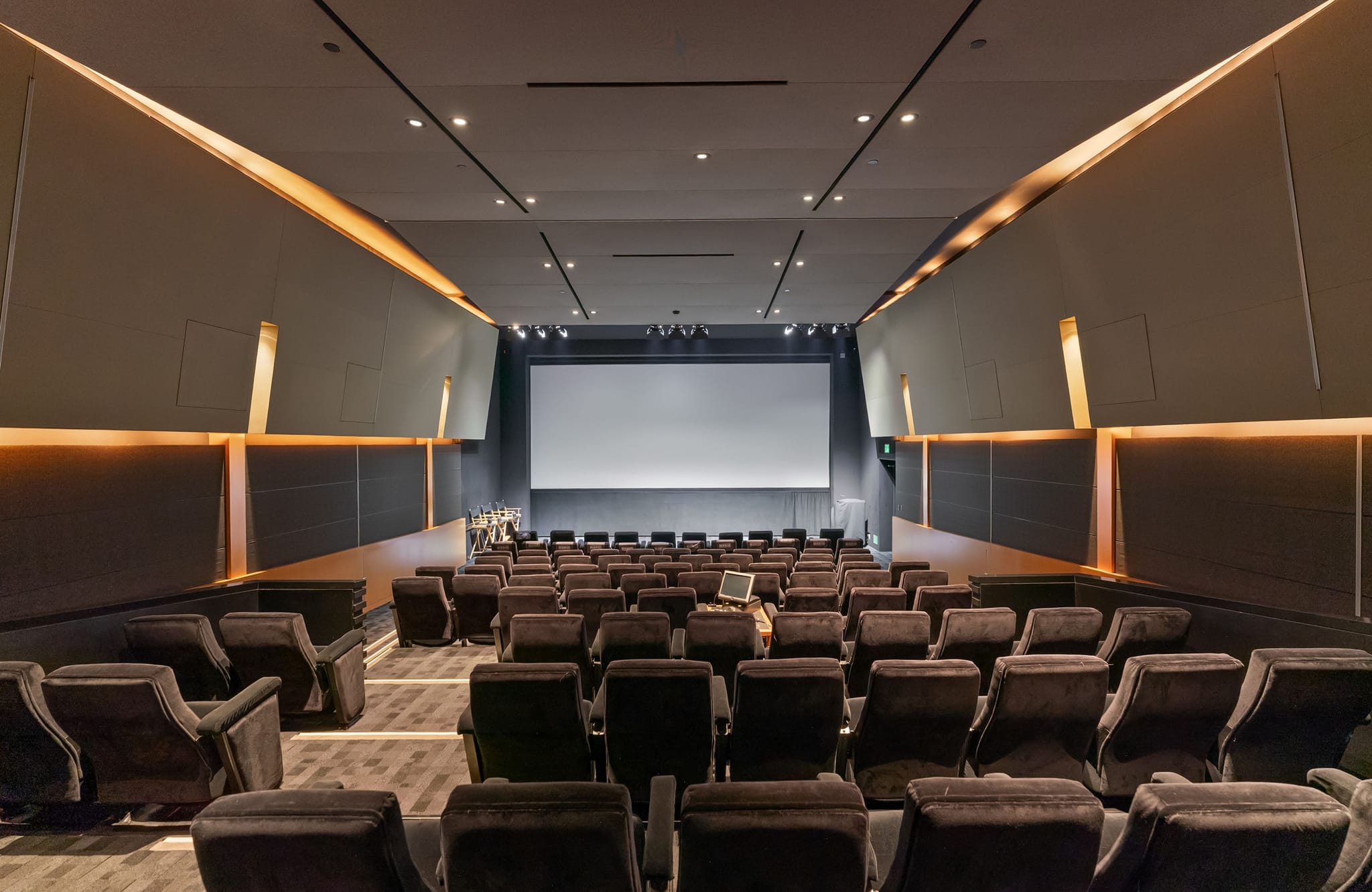 An empty, modern cinema with rows of dark, plush seating, a large screen, and dramatic orange accent lighting on the walls.