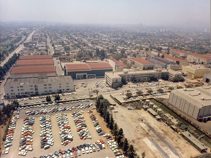 Aerial view of a sprawling studio lot with numerous buildings and parking lots packed with vintage cars, surrounded by a hazy city.