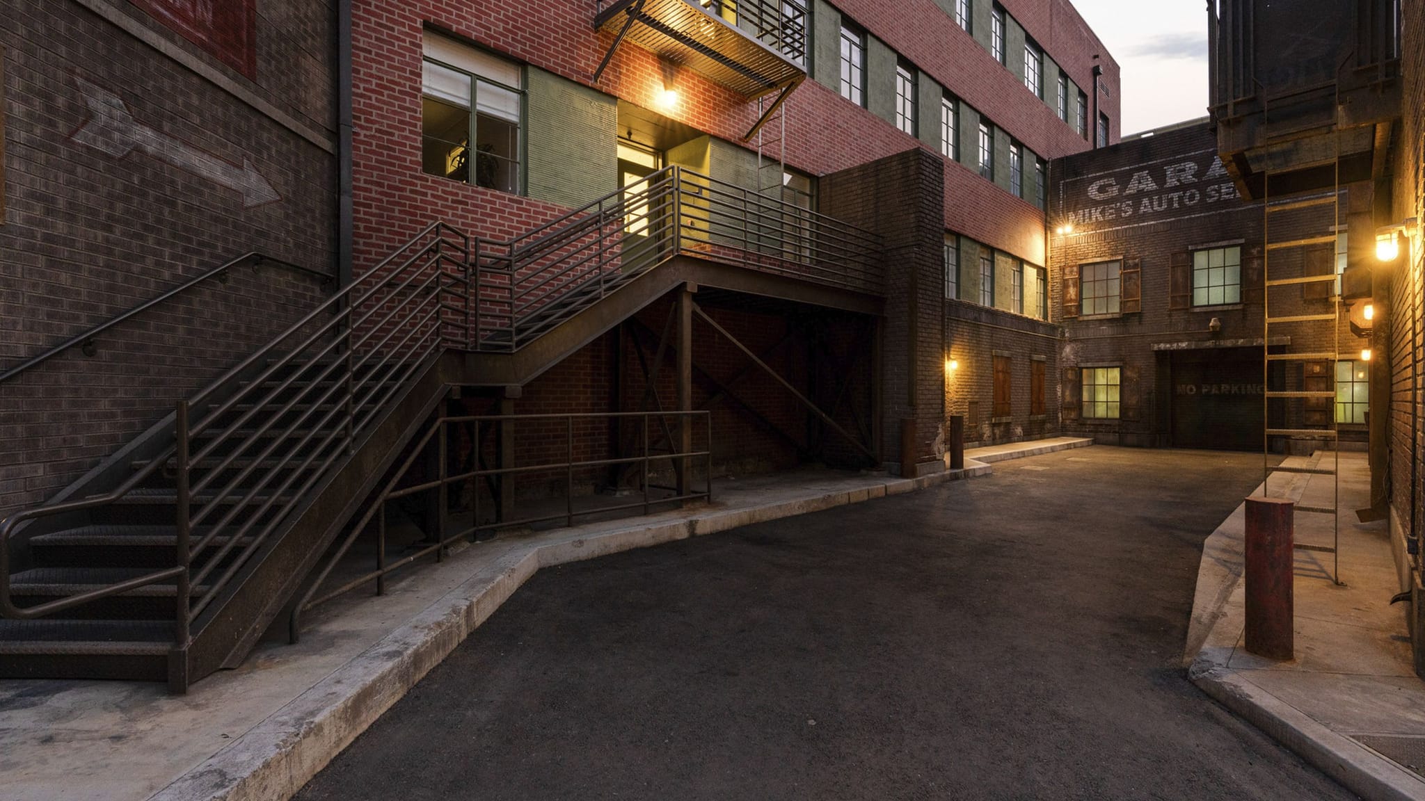 Empty brick alleyway with metal stairs and warm lights at dusk.