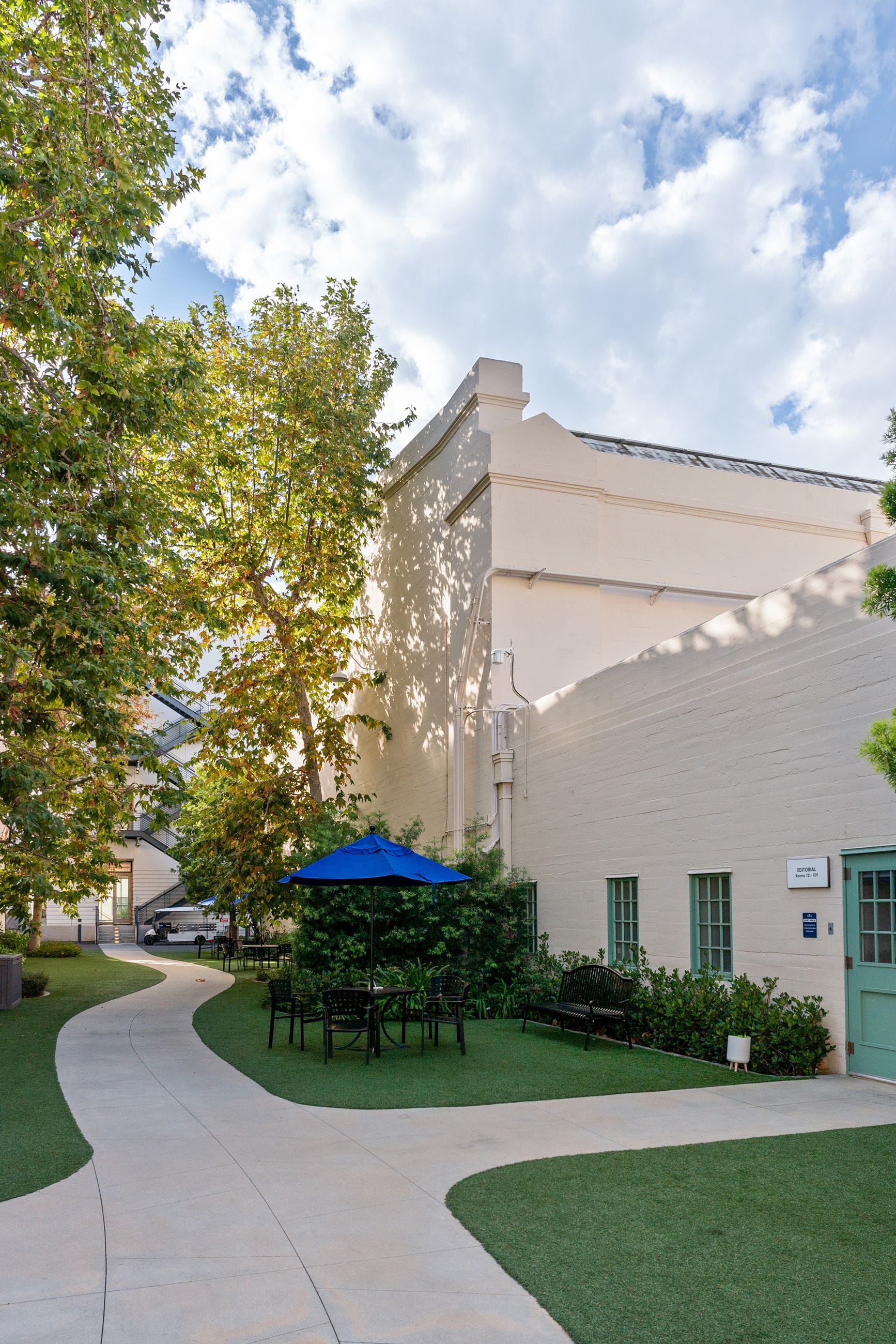 A courtyard with a winding concrete path, artificial grass, a blue umbrella over outdoor seating, and a beige building surrounded by trees under a cloudy sky.