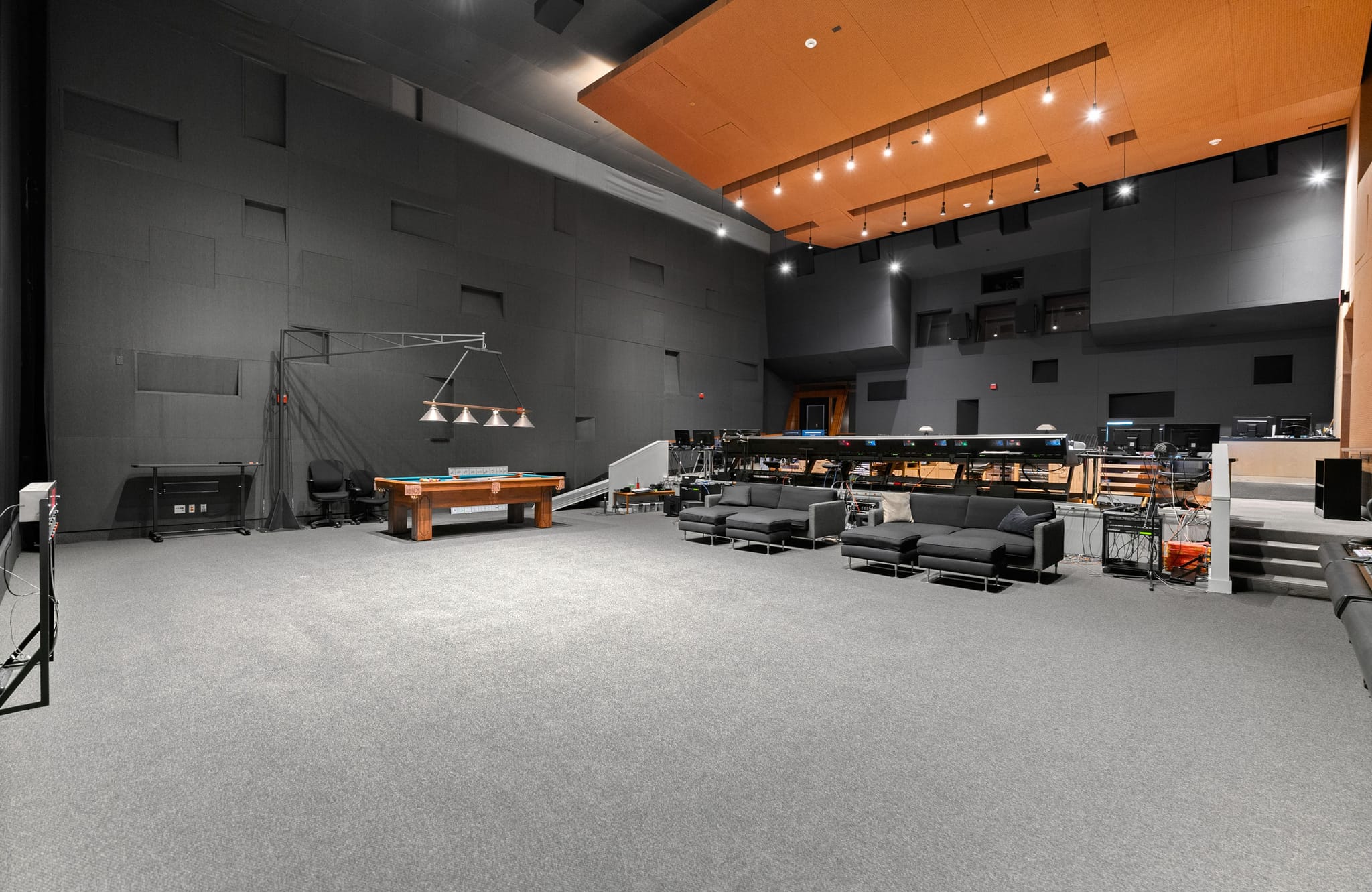 A large studio space featuring a pool table, multiple dark sofas, and audio production desks.