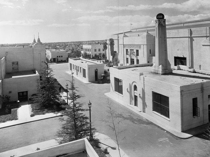 Black and white photo of an architectural complex with white buildings, paved roads, and trees under a cloudy sky.