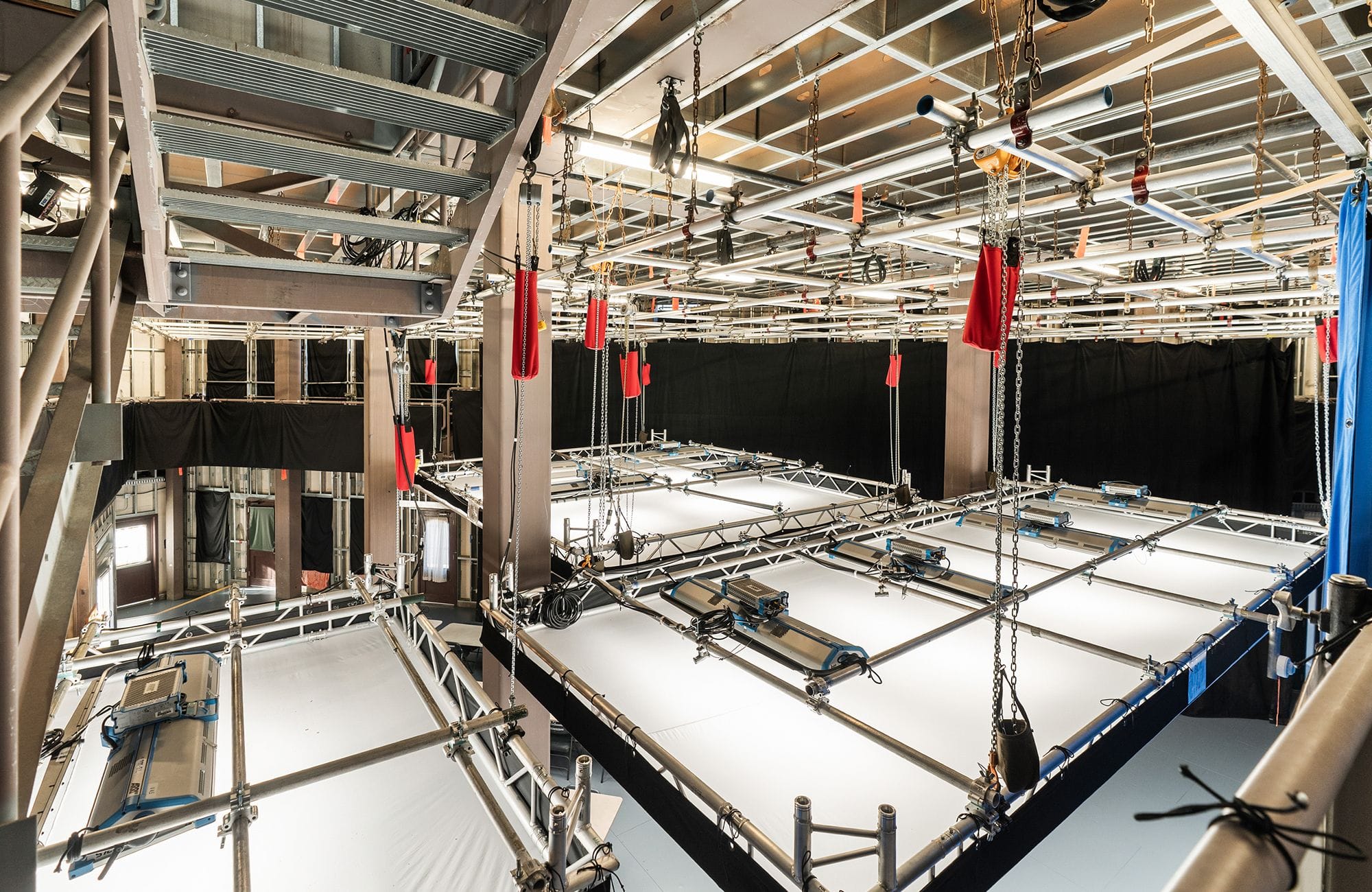 A large, high-tech studio with overhead metal scaffolding, hanging rigging equipment including red elements, and large white modular platforms.