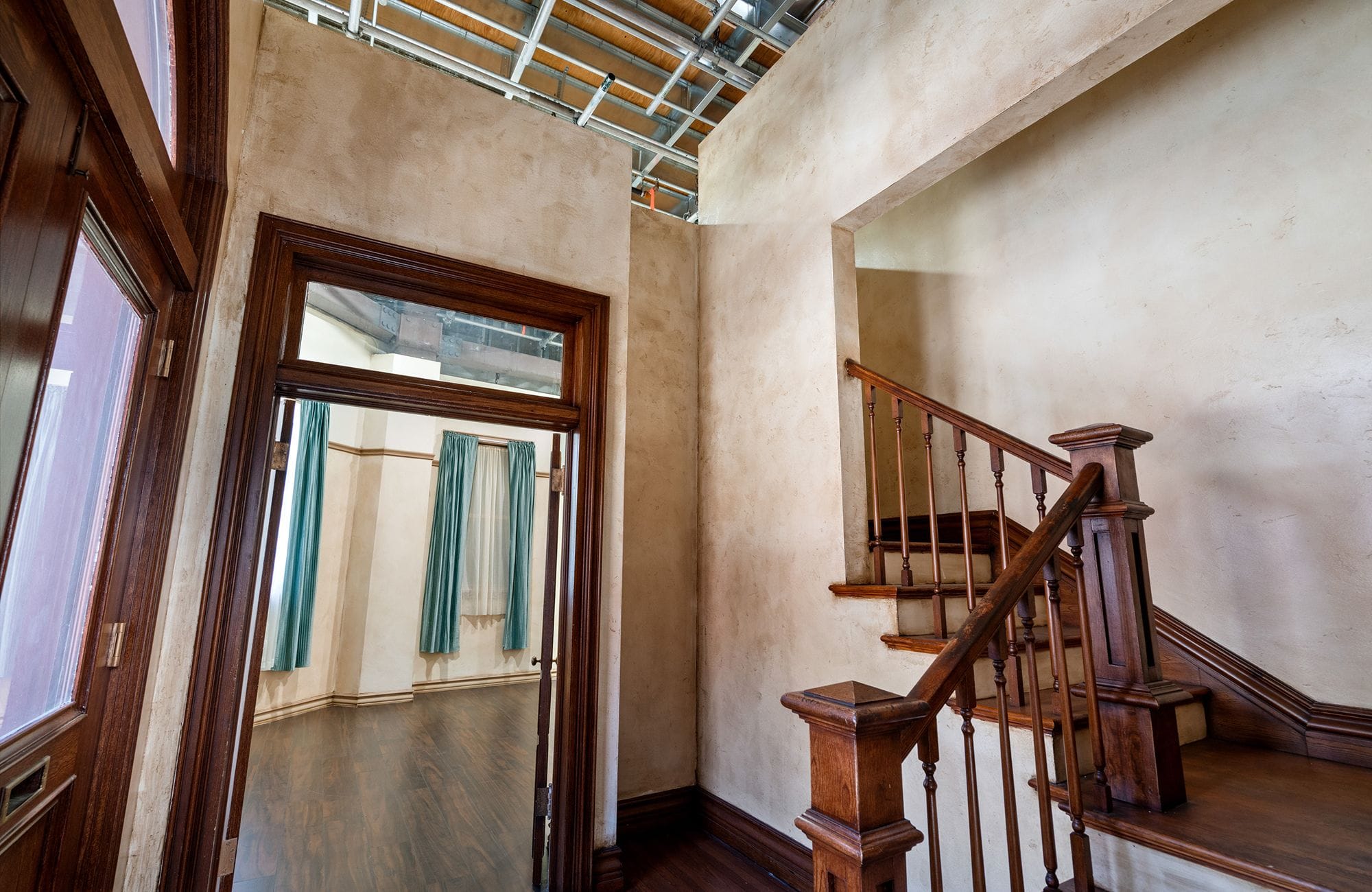 Interior set with a dark wood staircase, a doorway revealing a room with teal curtains, and an exposed ceiling structure.