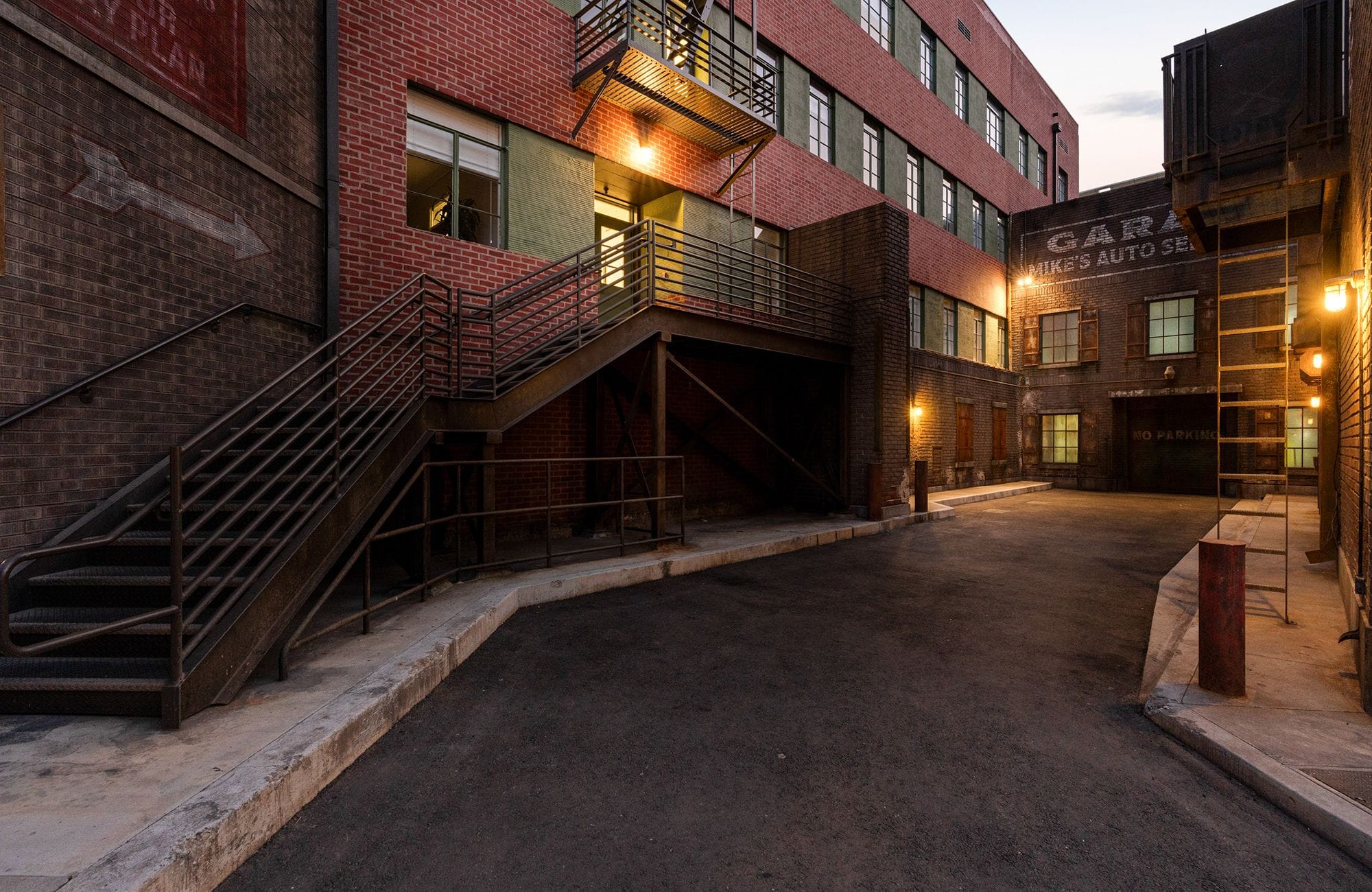 Industrial alleyway at dusk, with brick buildings, metal stairs, a paved path, and warm lights illuminating windows and a 'Garage' sign.
