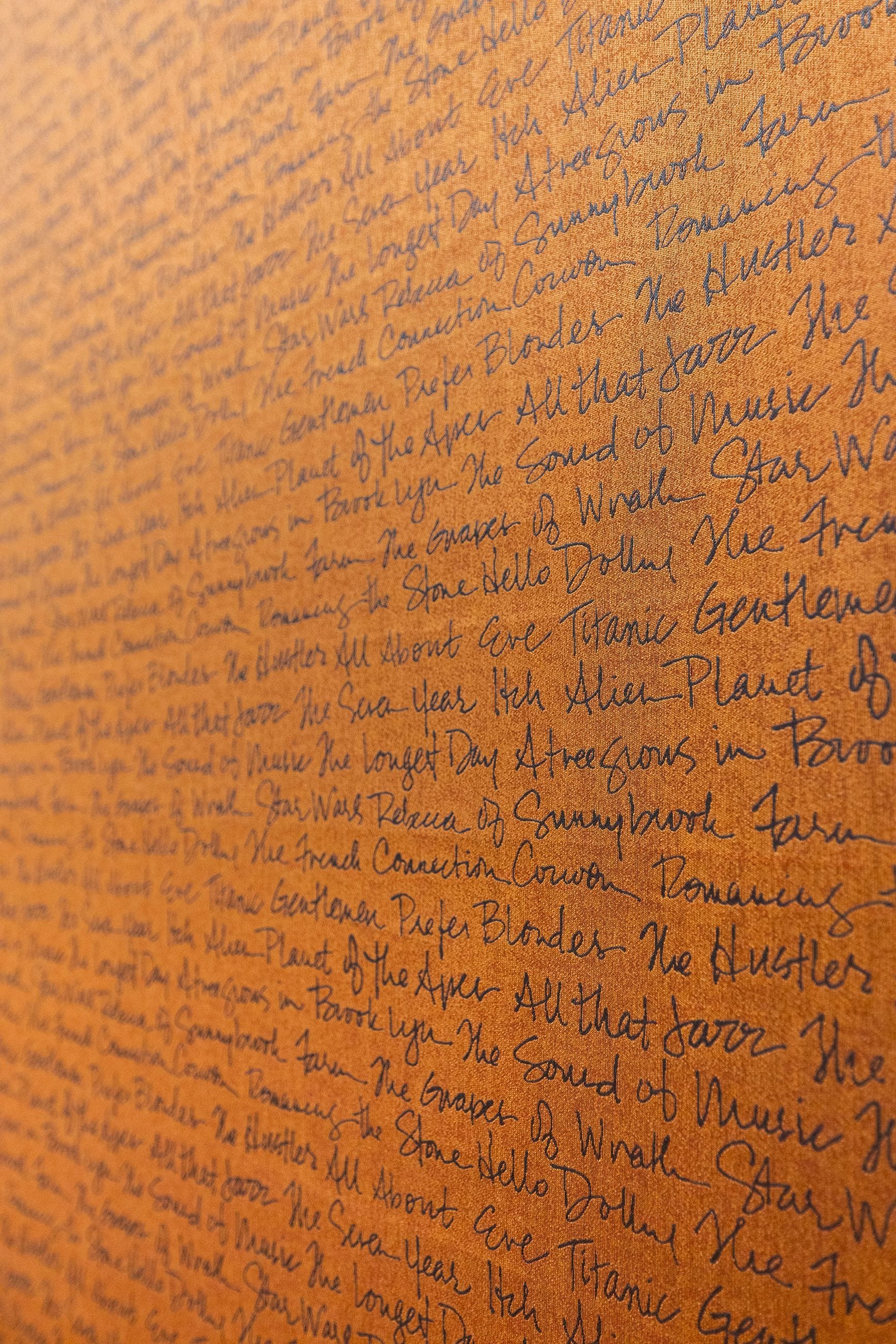 A close-up of dark, handwritten movie titles densely covering an orange-brown paper, with some text blurred from the angled perspective.