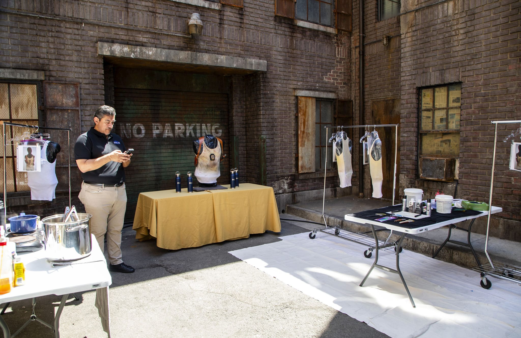 A man looks at his phone in a brick alleyway with tables displaying clothing, a torso mannequin, and supplies, against a garage door reading "NO PARKING."