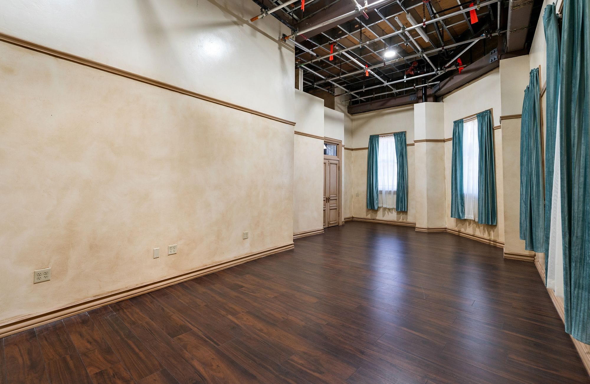 An empty studio room with dark wood floors, cream and beige walls, teal curtains on windows, and an exposed grid ceiling.