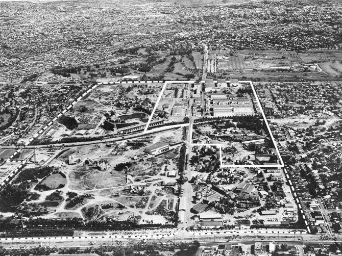 Black and white aerial view of a sprawling urban area with a large amusement park or fairgrounds outlined by a dashed white line.