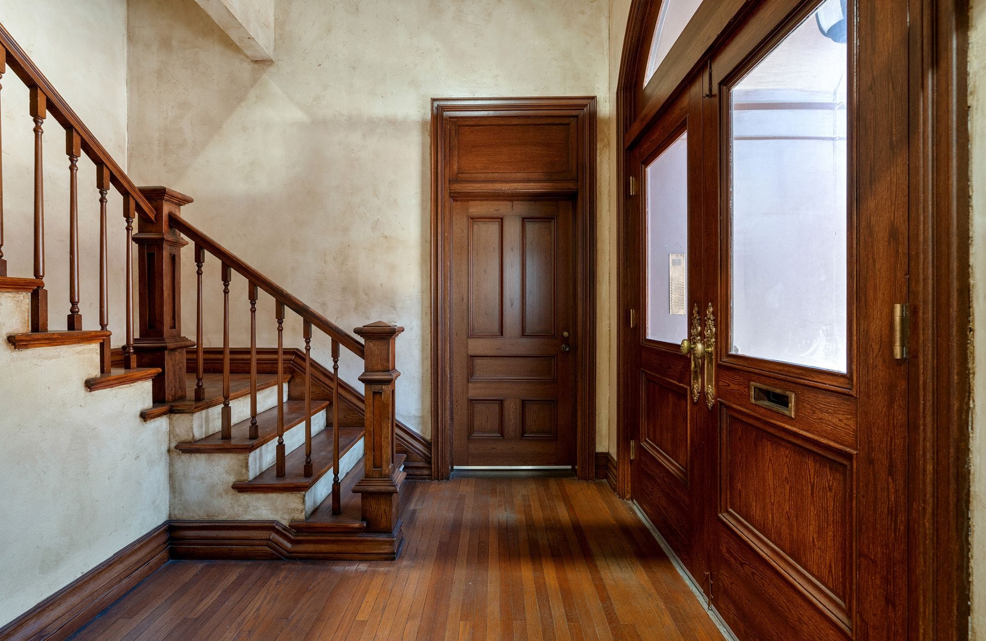 A hallway with a wooden staircase, two dark wood doors, and hardwood floors.