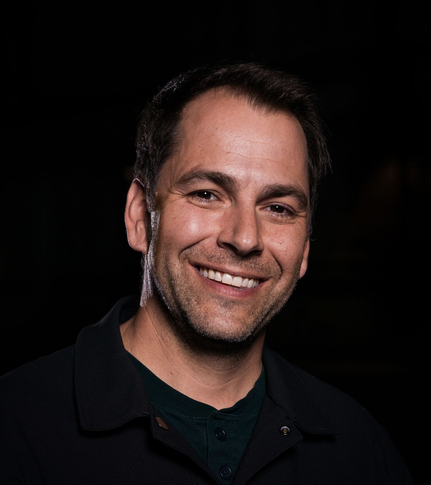 A smiling man with dark hair and stubble, wearing a dark collared shirt.