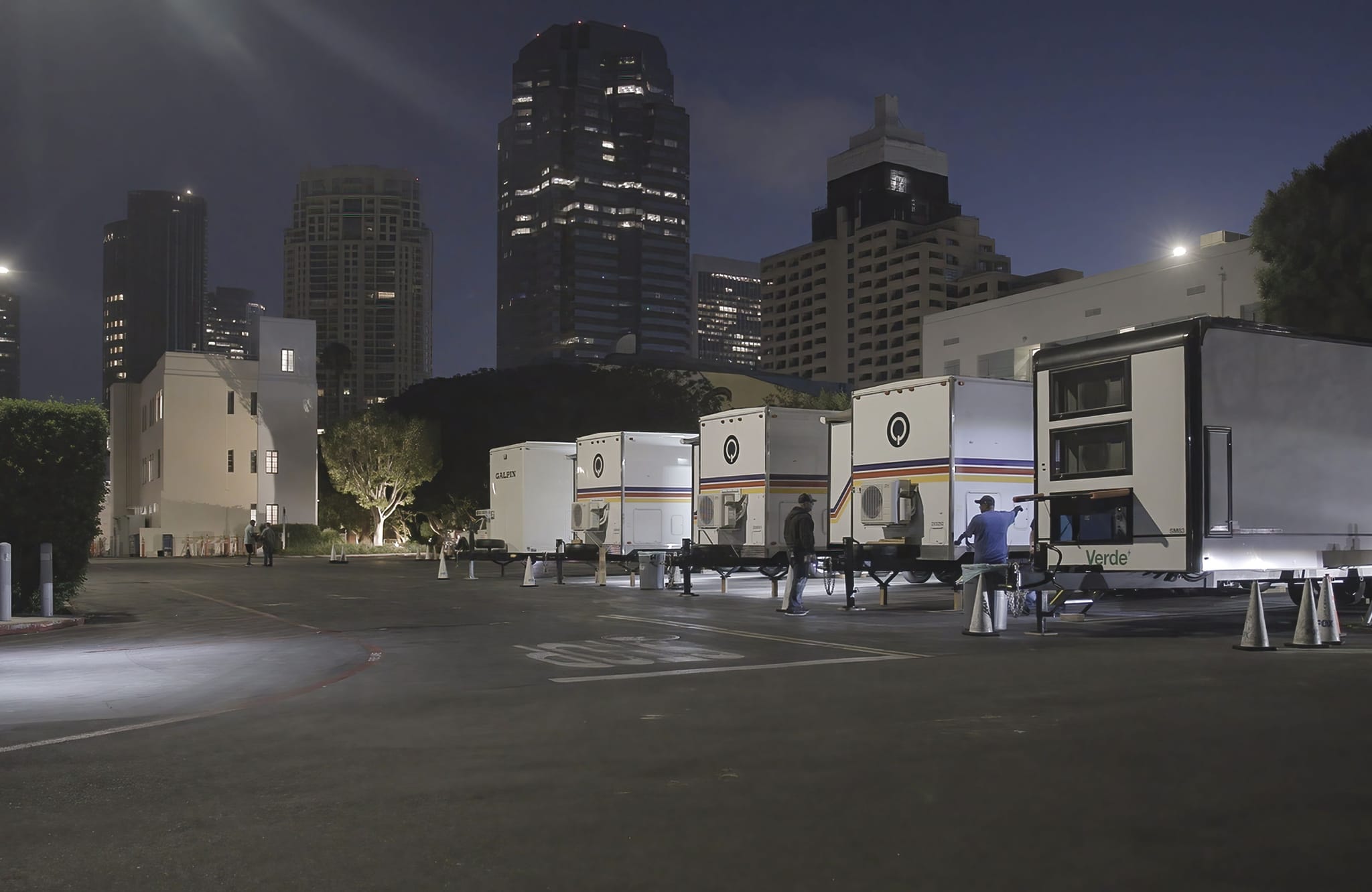 White portable units are parked in a lot at night, with city buildings illuminated in the background.