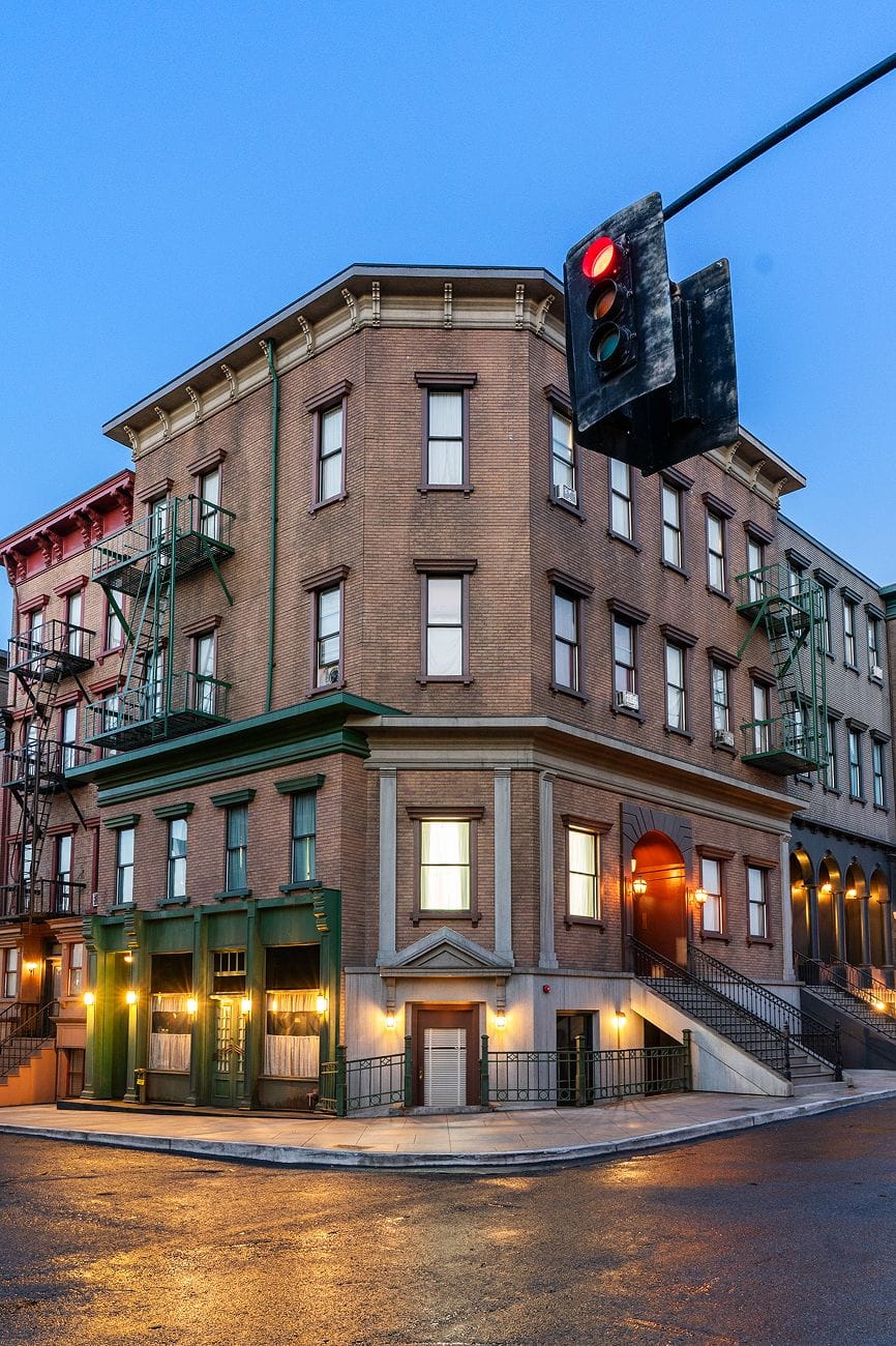 A brick corner building with illuminated windows, fire escapes, a red traffic light, and reflections on wet pavement at dusk.