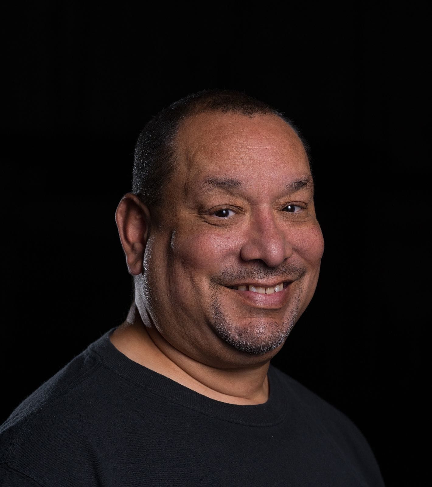 A smiling man with a goatee and short hair wearing a black t-shirt against a dark background.